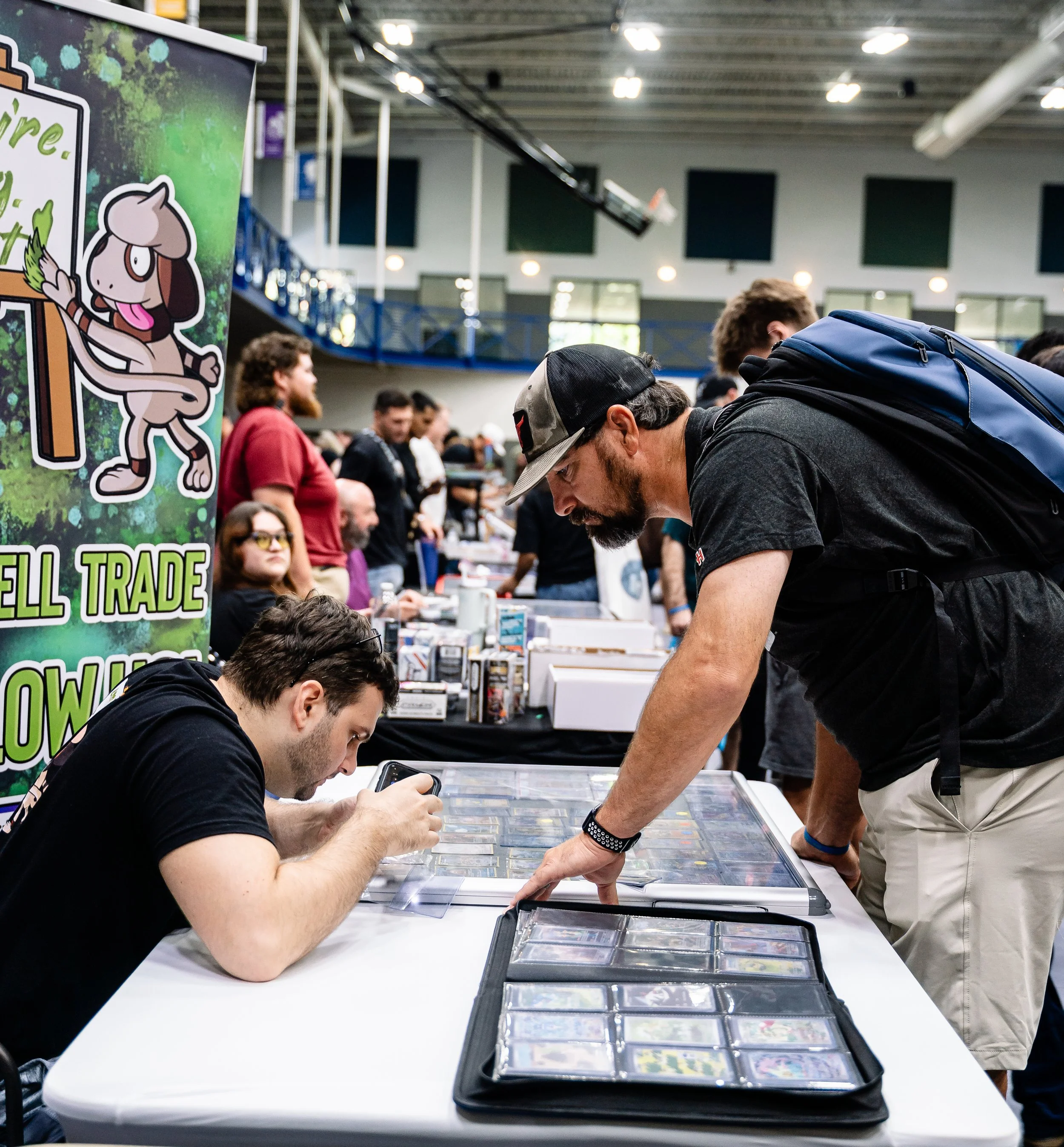 A group of people at a trading event or convention, gathered around a table with trading cards and collectibles, with a banner promoting trading.