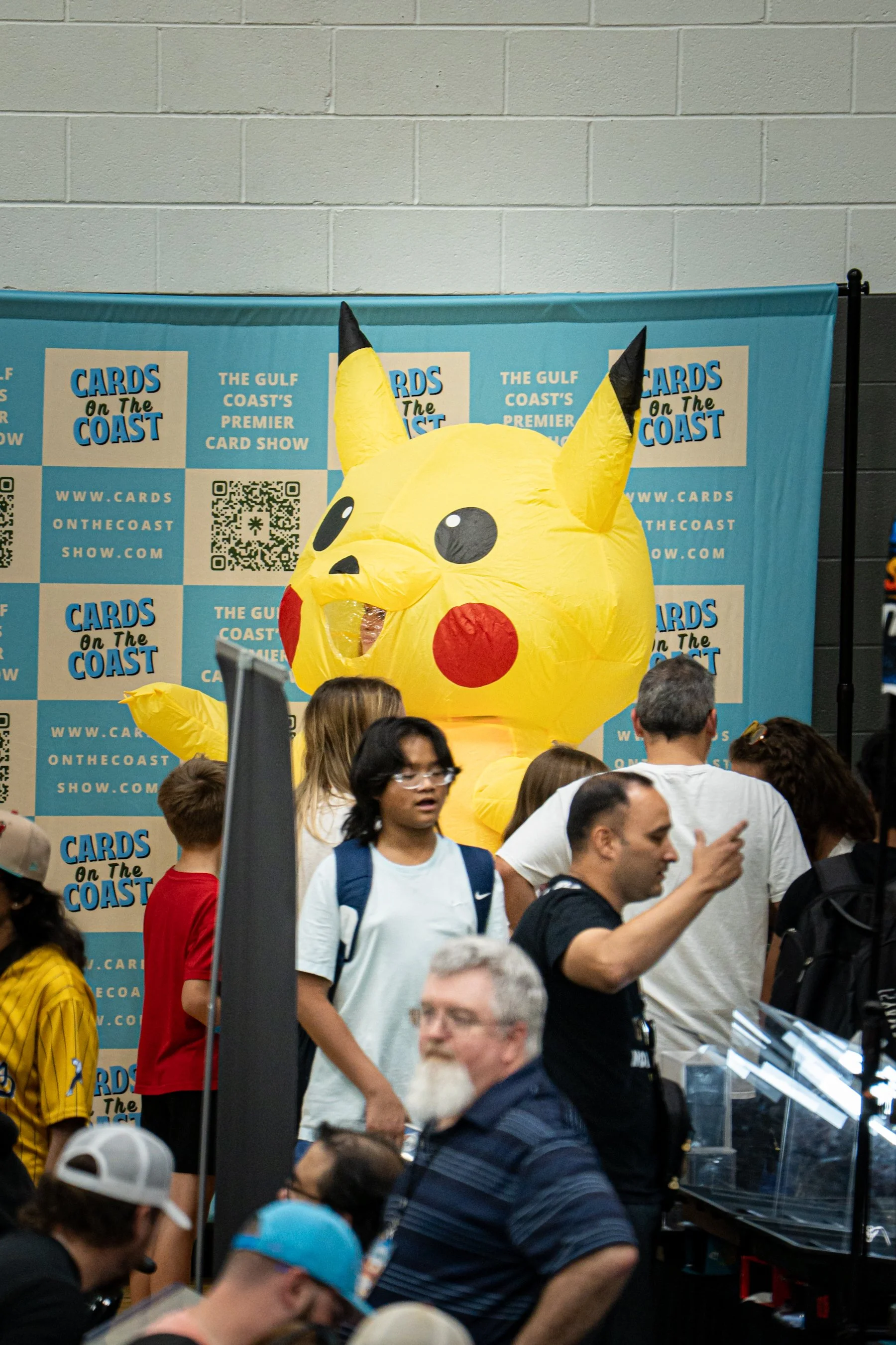A large Pikachu mascot costume at the Cards on the Coast event, with a blue backdrop displaying event information and people gathered in front.