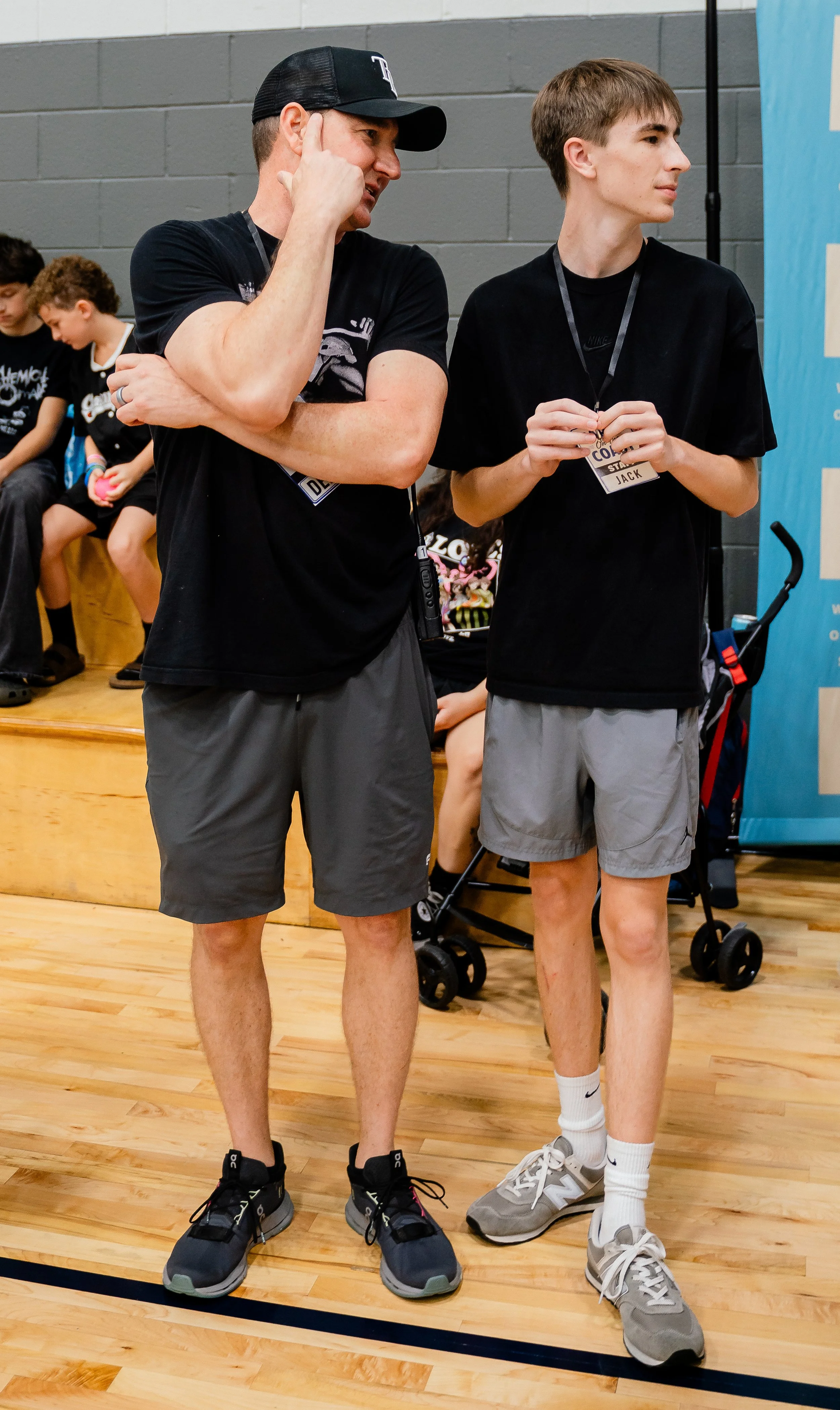 Two young men standing in a gymnasium, engaged in conversation. The man on the left is wearing a black Cap, black t-shirt, gray shorts, and running shoes; he has a listen on his neck and a name badge hanging from a lanyard. The young man on the right