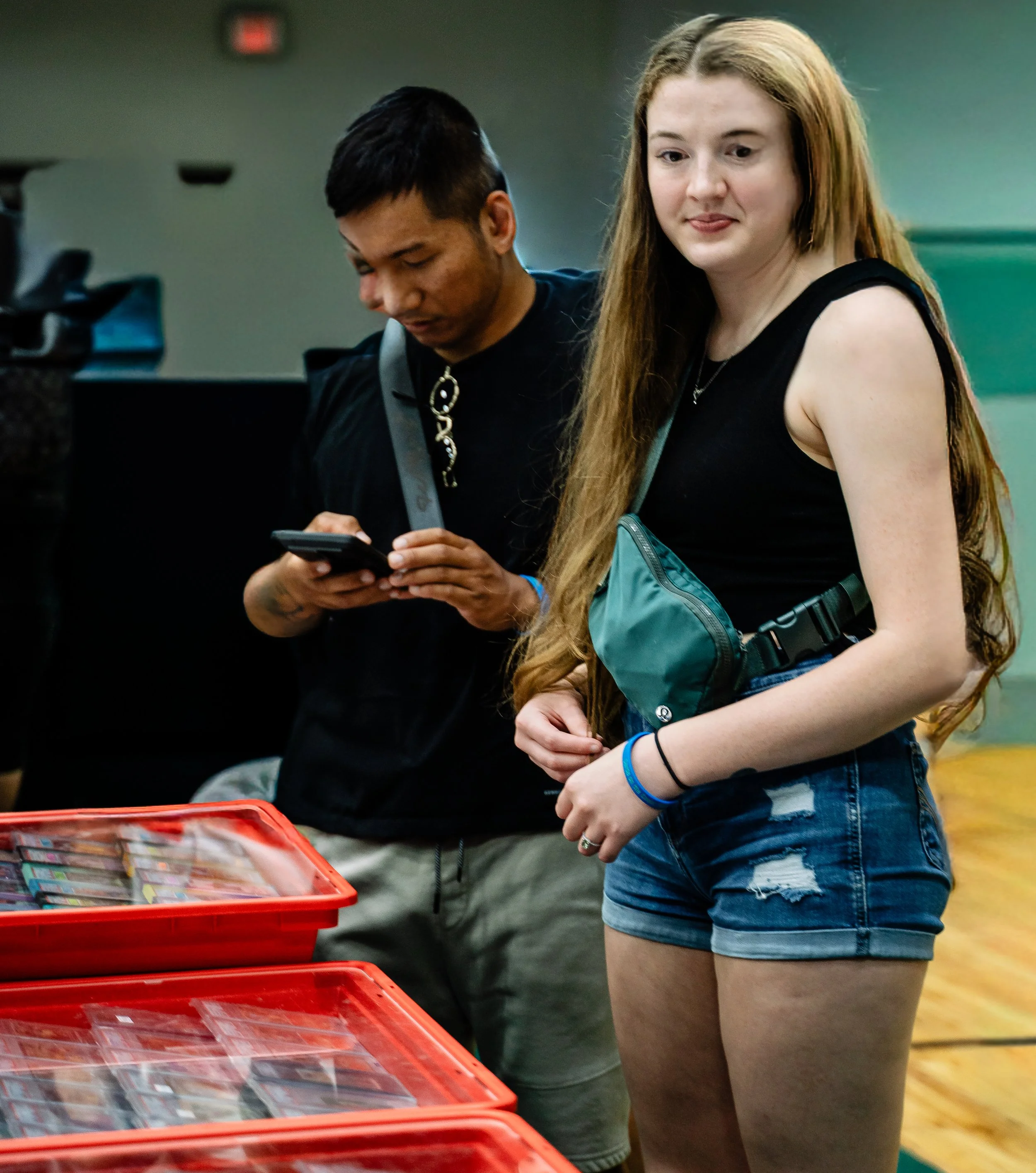 Two young women at a collectible card display table, one using a smartphone and the other with long hair and casual clothing, in an indoor setting.