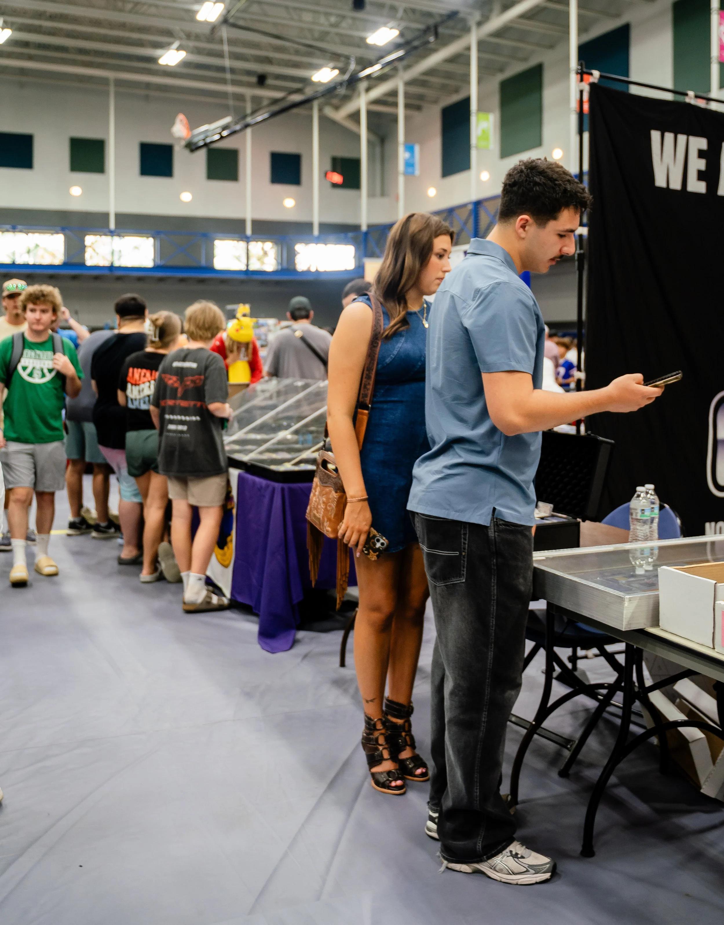 People standing in line at an indoor event, with a woman in high-heeled sandals and a man in casual sneakers looking at a phone, various others in line behind them.