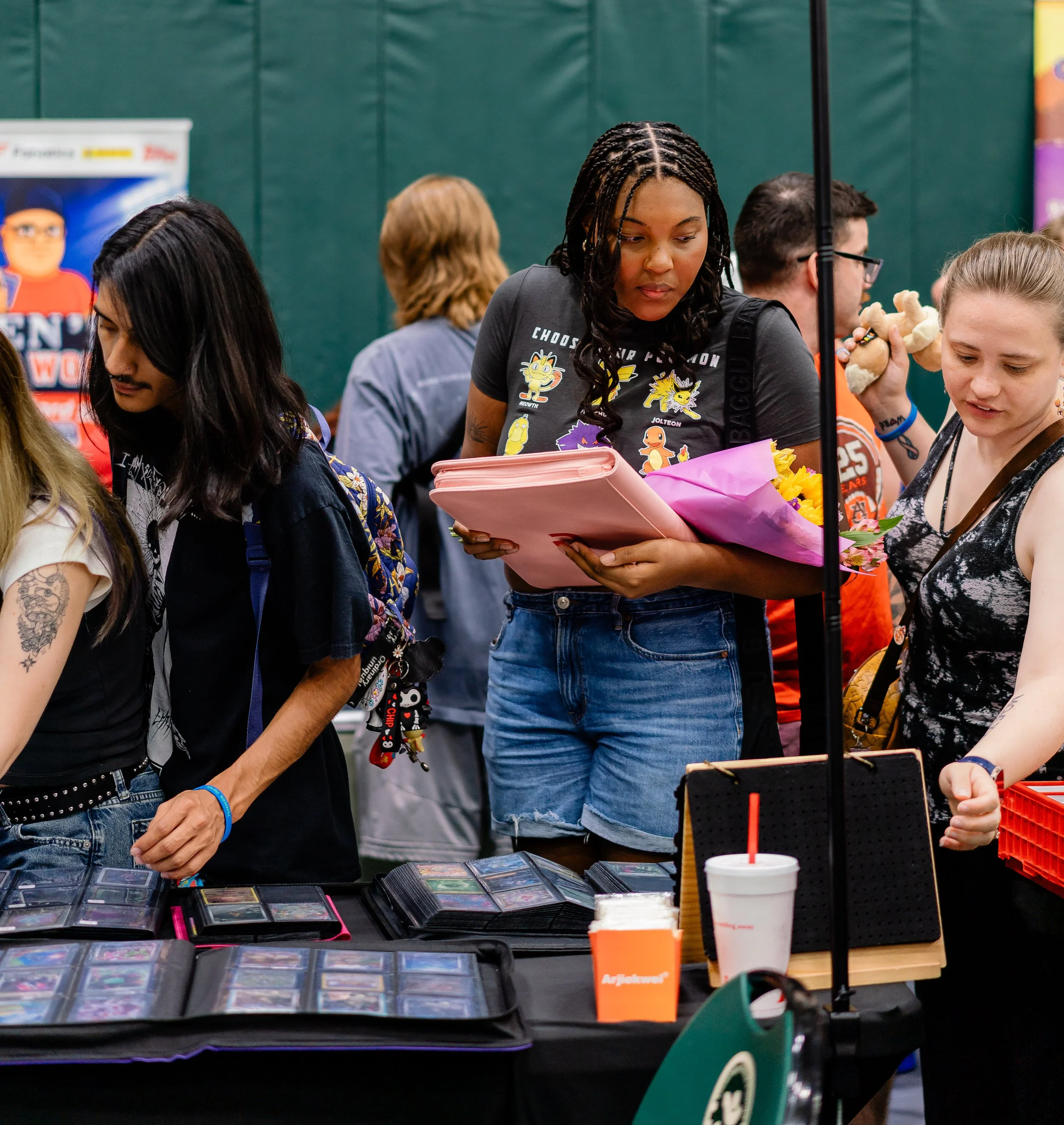 A group of people gathered around a table at a convention or event, looking at trading cards and merchandise. There are several card binders open on the table, displaying multiple trading cards. A woman in the center wearing a black T-shirt with cart