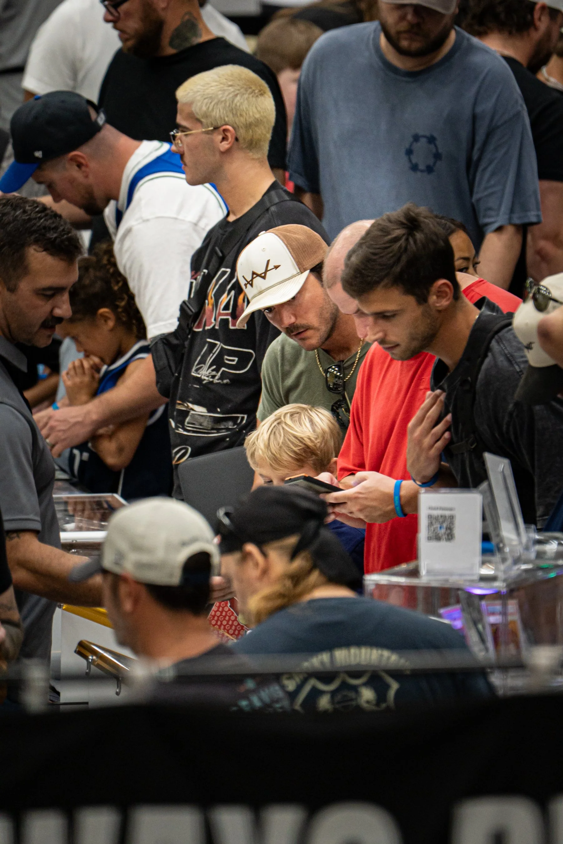 Crowd of people gathered at a table, some looking at electronic devices, in an indoor setting, likely a convention or event.