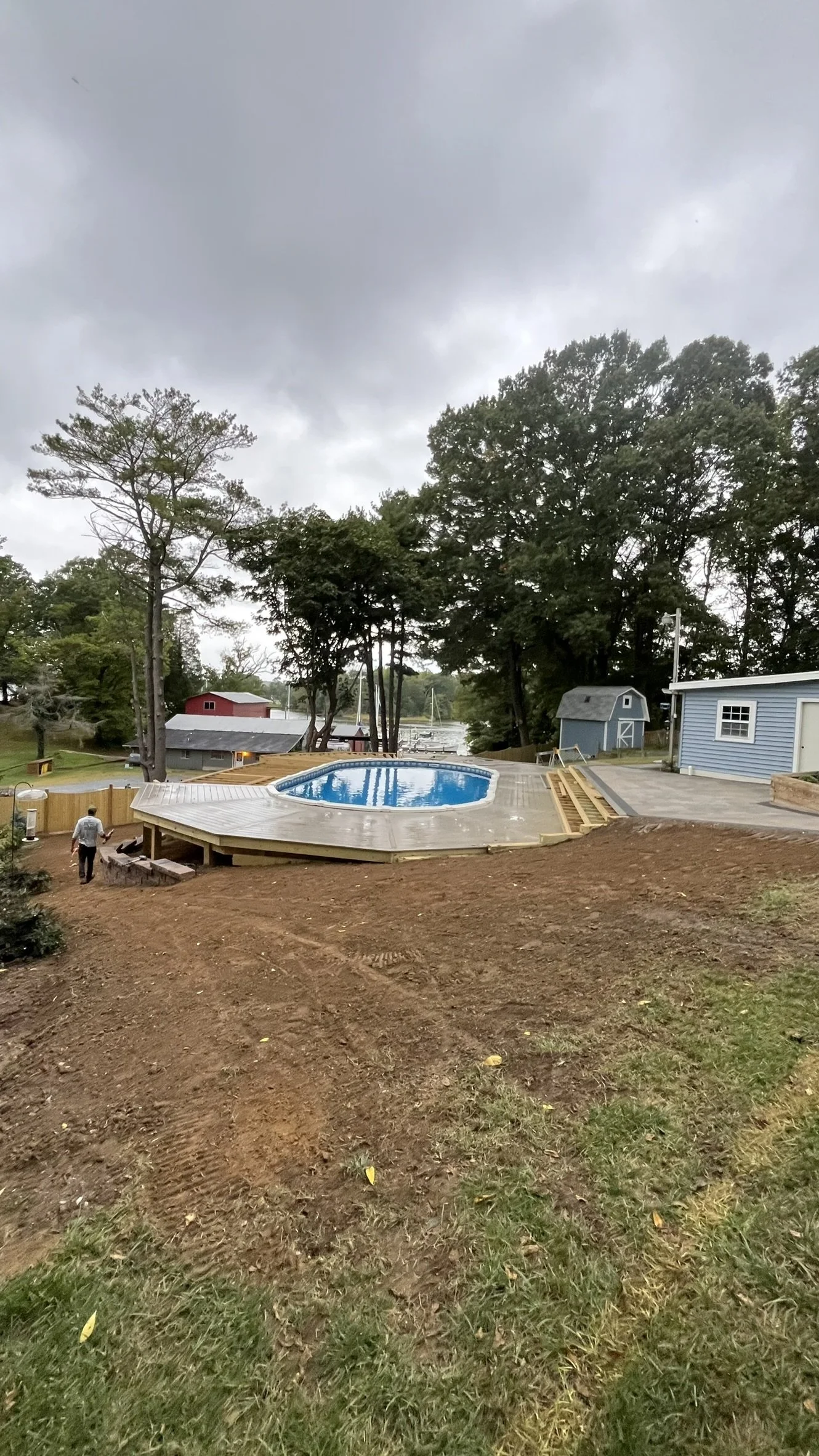 A newly built wooden deck surrounds a small above-ground swimming pool situated in a backyard. The yard has some green grass and areas of bare, brown soil. There are trees, a blue shed, and a blue house in the background. The sky is overcast with dark gray clouds.