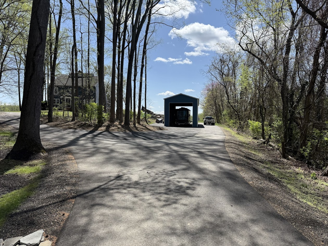 A paved driveway leading to a small blue garage with an open door, housing a parked recreational vehicle. Two cars are parked outside, one on each side of the garage. Tall trees line both sides of the driveway, casting shadows on the ground, with a partly cloudy sky overhead.