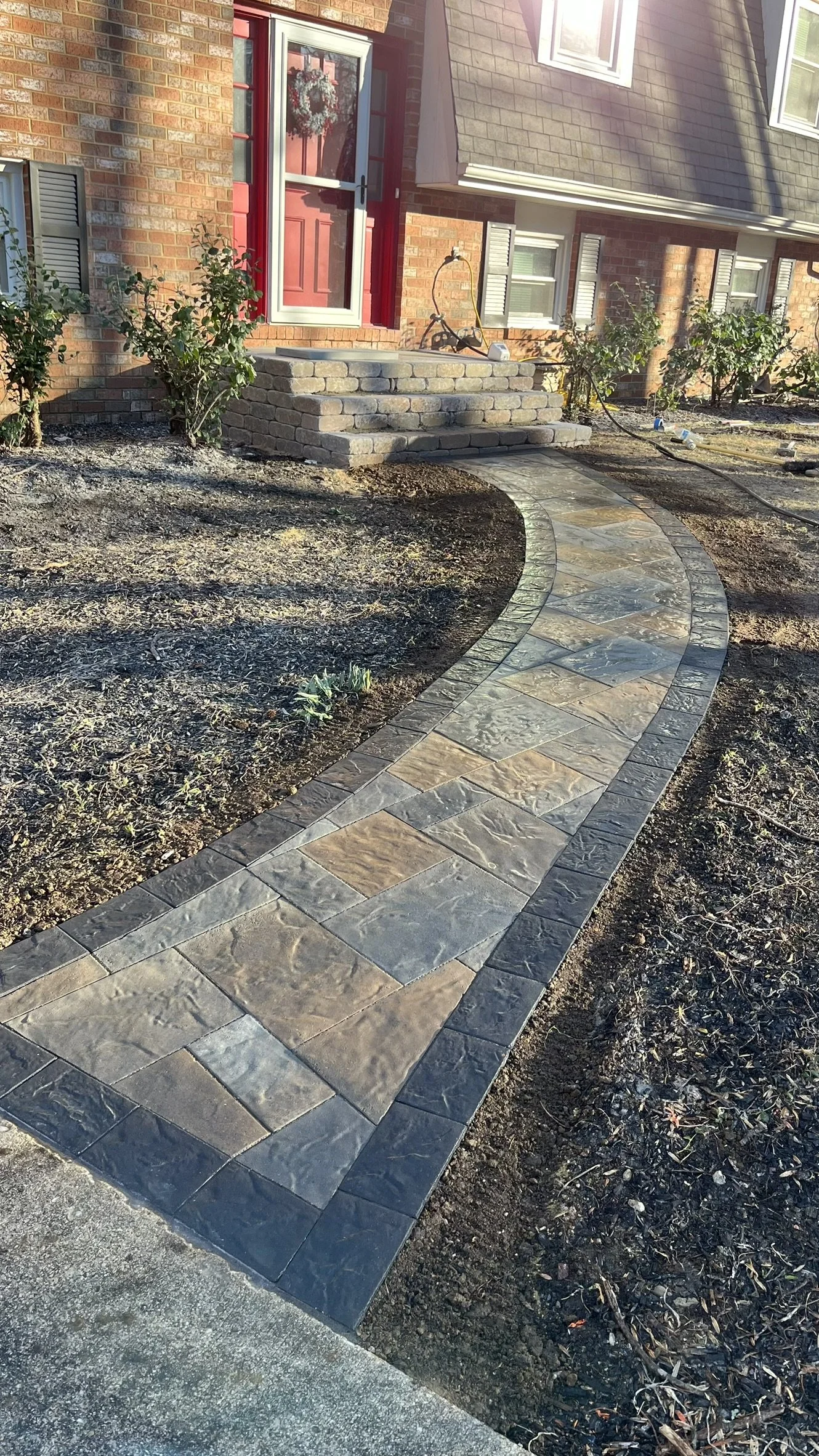 Curved stone pathway leading to front steps of a brick house with a red door and porch steps, surrounded by garden beds with small shrubs.