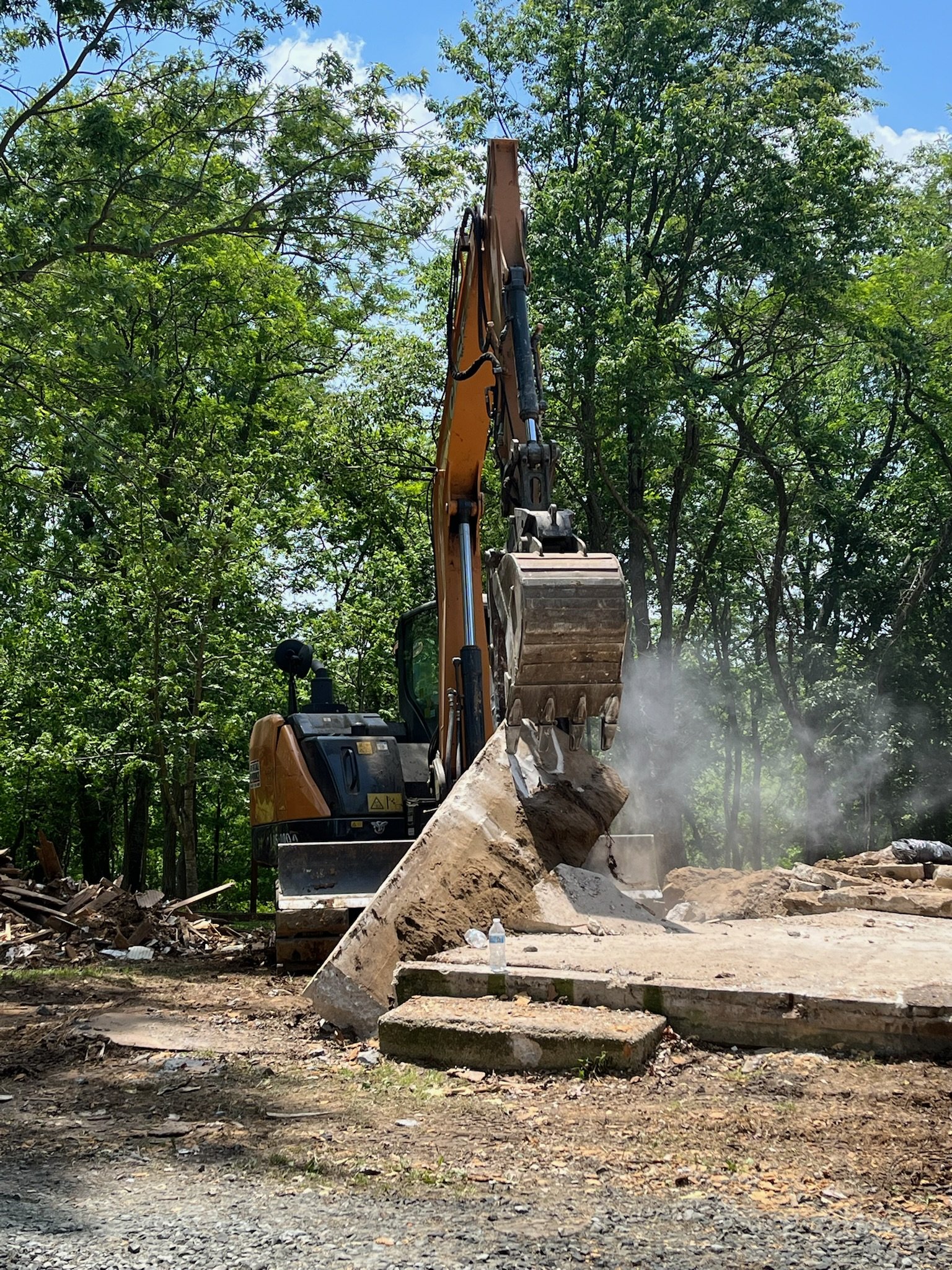 A large excavator demolishing a concrete structure with debris and dust, surrounded by green trees and a blue sky.