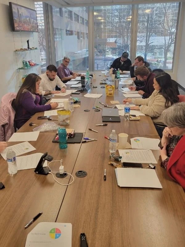 A group of people sitting around a long conference table in a meeting room, working on laptops and reading documents, with large windows showing a cityscape outside.