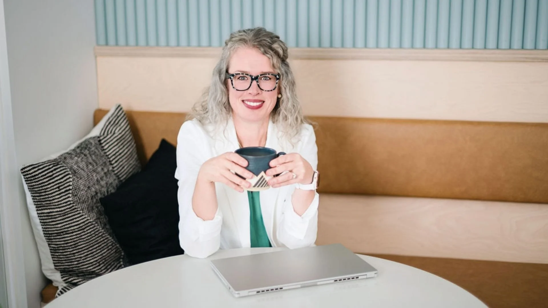 Heather Beck, A smiling woman with glasses and curly blonde hair sitting at a table, holding a mug. A closed laptop is in front of her on the table, and black-and-white pillows are on a bench beside her.
