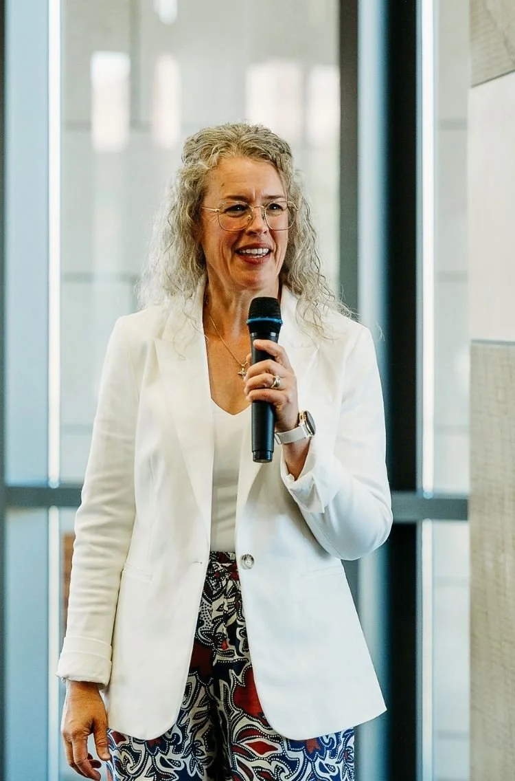 Heather Beck, A woman with curly gray hair and glasses holding a microphone, speaking indoors.