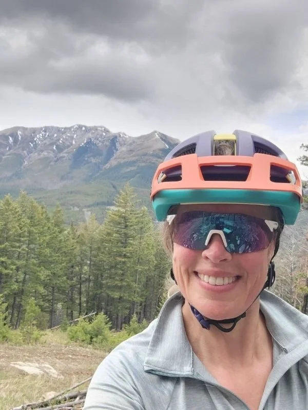 Woman wearing a colorful bike helmet and sunglasses, smiling in front of mountains and forested landscape.