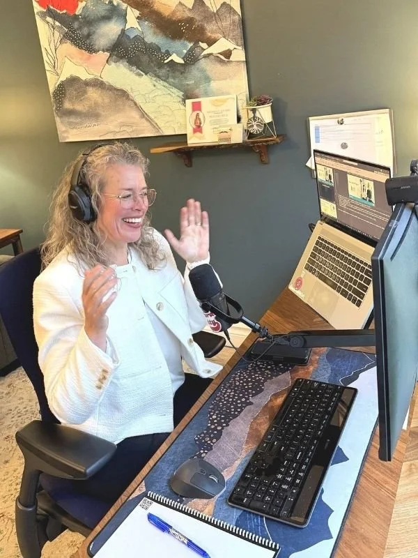 A woman with curly gray hair, wearing glasses and a white jacket, is seated at a desk with a microphone, computer monitor, and laptop, smiling and waving during a video recording or virtual meeting.
