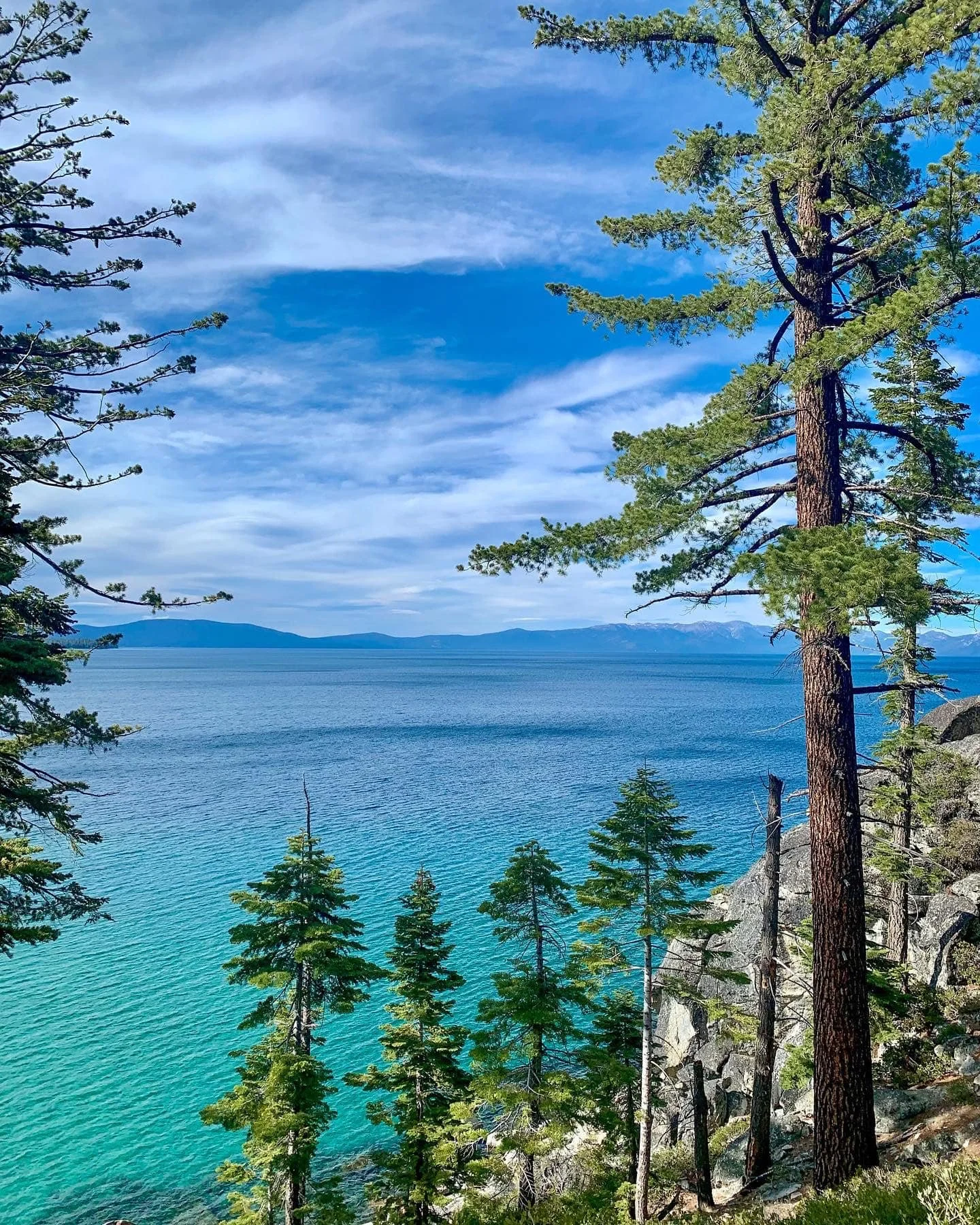 Serene lakeside view with green pine trees on a rocky slope and a calm blue lake extending to distant mountains under a partly cloudy sky.