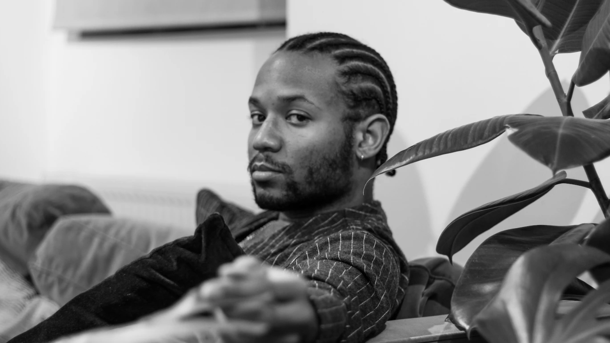 Black and white portrait of a person with braids, sitting on a couch next to a potted plant.
