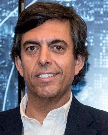 A man with dark hair wearing a white collared shirt and a dark jacket, smiling against a cityscape background.