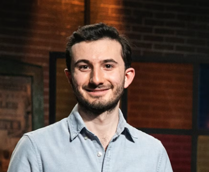 A smiling man with dark hair and a beard, wearing a light blue button-up shirt, standing indoors against a background of a brick wall and framed artwork.