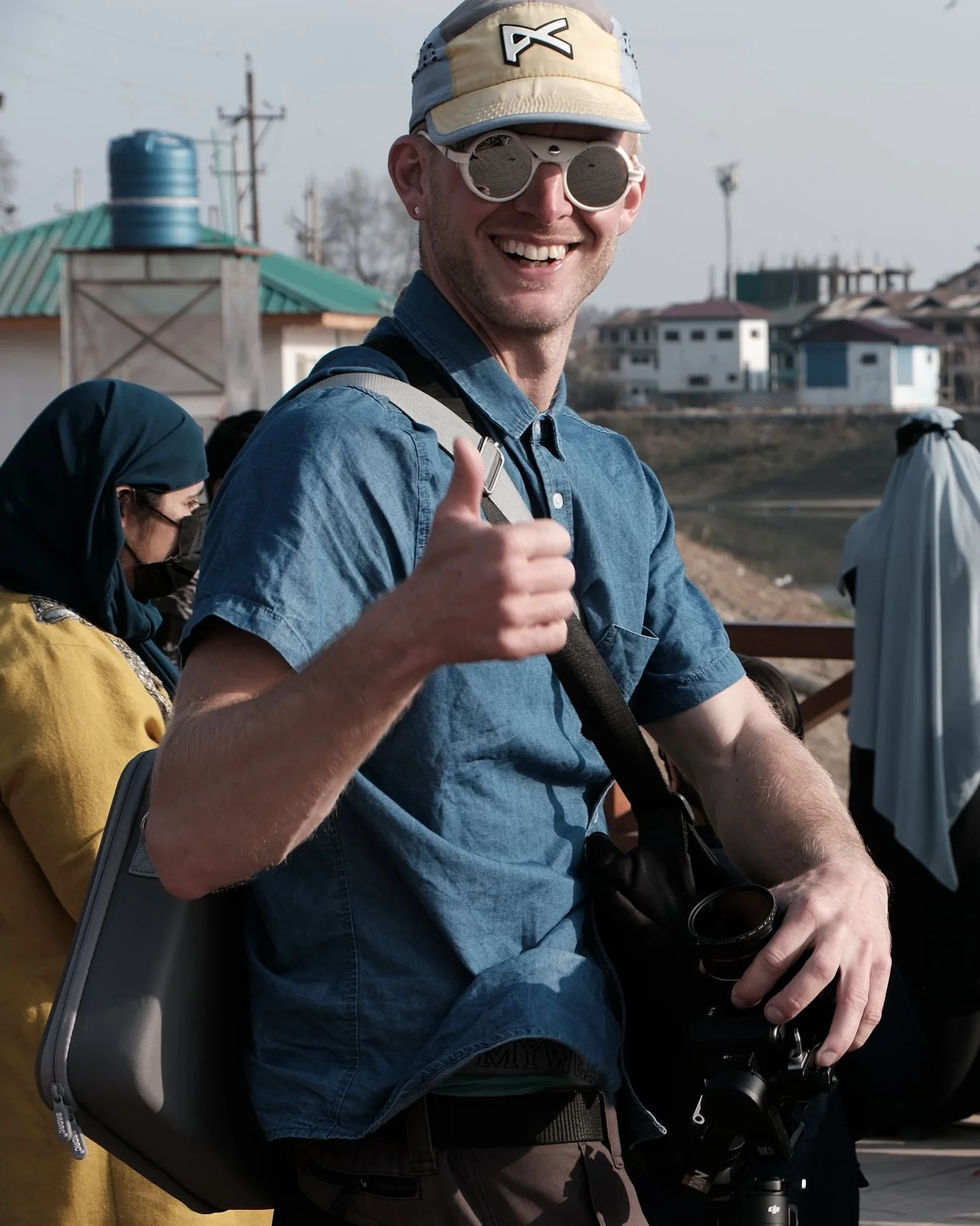 Harald Edlund, renowned outdoor sports videographer and documentary filmmaker based in Switzerland. sunglasses, and a blue denim shirt giving a thumbs-up while holding a camera, outdoors on a cloudy day with people and buildings in the background.