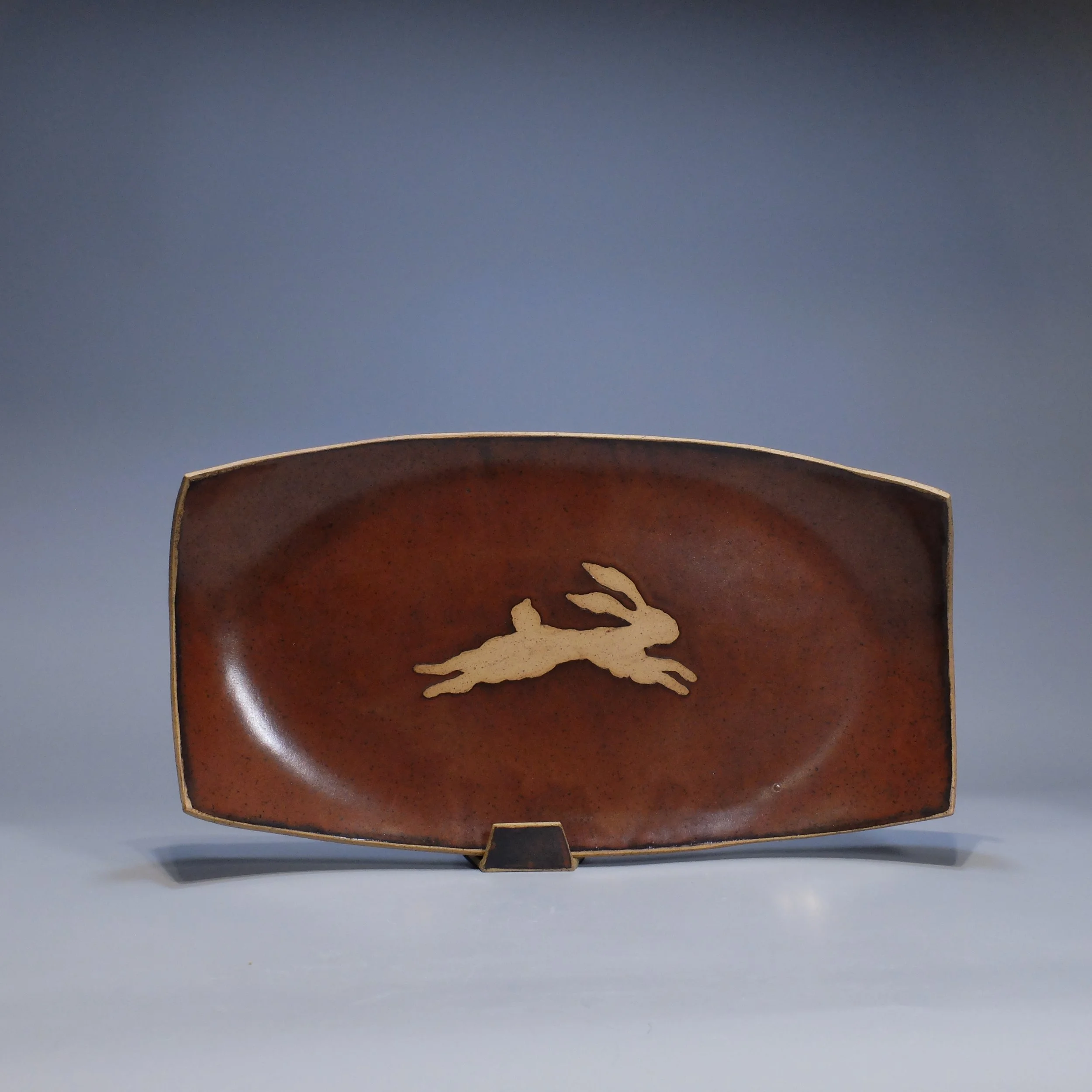 Platter, 2026. Speckled stoneware, red iron oxide glaze, leaping hare motif. 17.25"L, 8.75"W, 2"D.