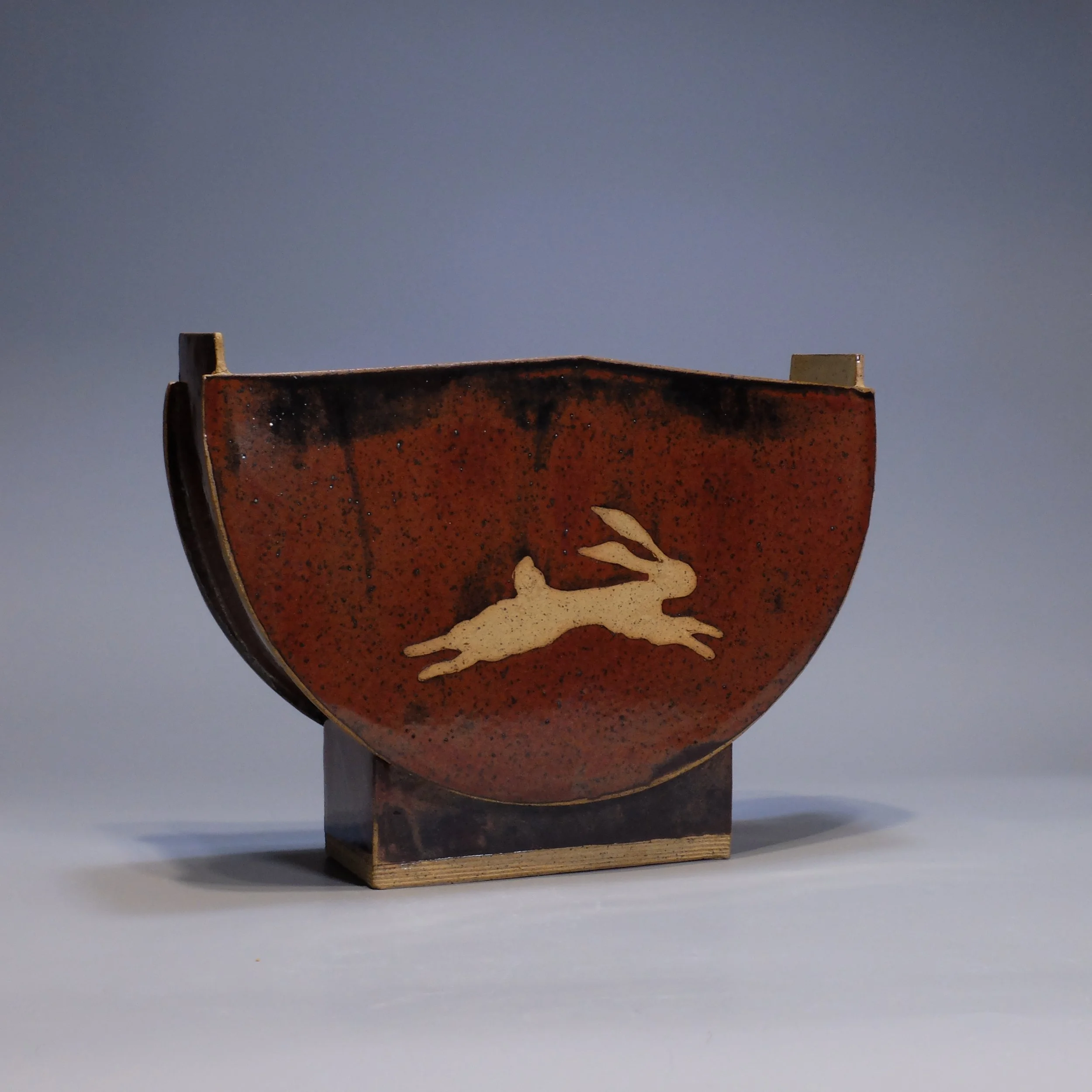 Half-Moon Vase, 2026. Speckled stoneware, red iron oxide glaze, leaping hare motif. 12.25"W, 8.25"T, 3.75"D