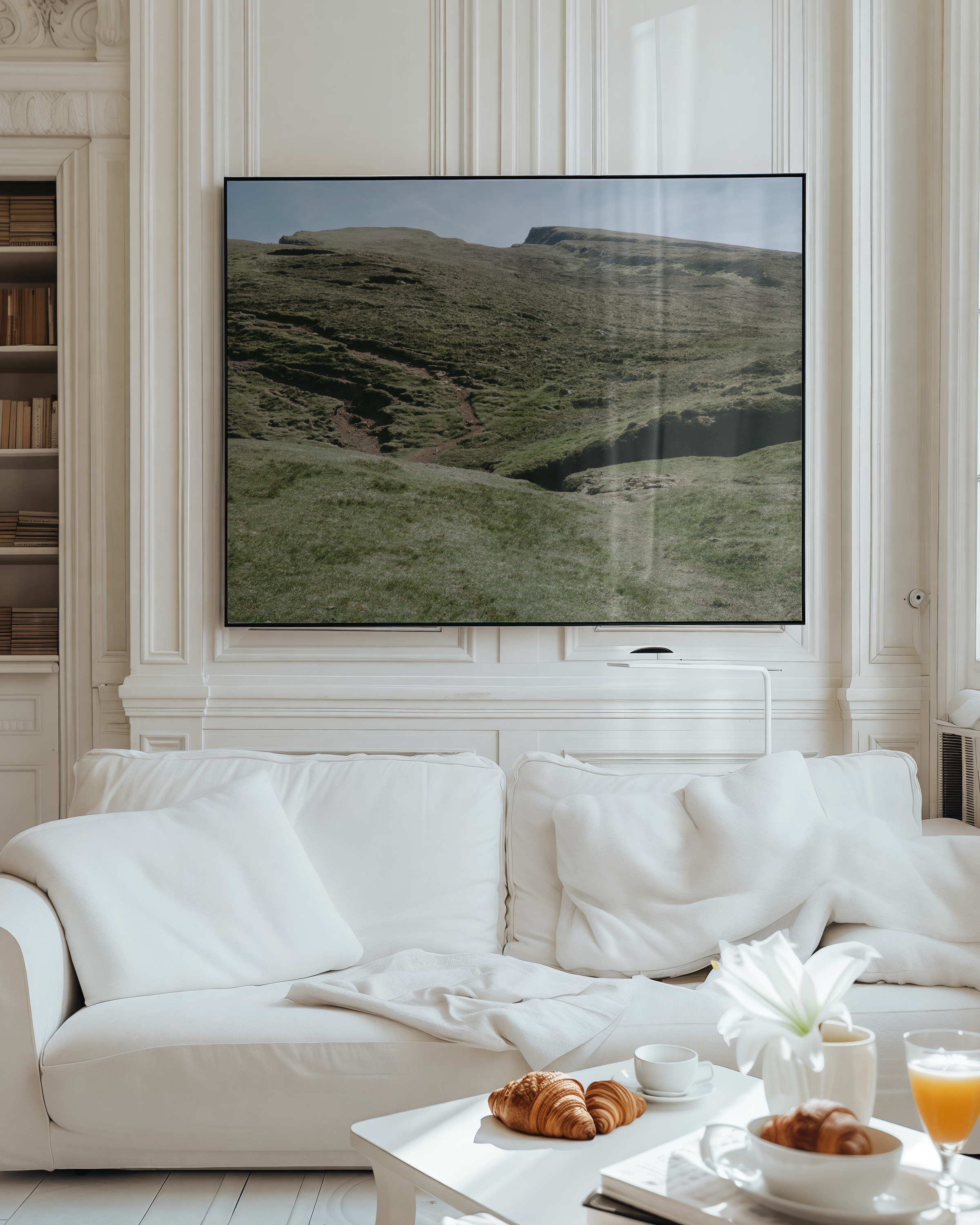 Living room with a large flat-screen TV showing a mountainous landscape, white sofa with pillows, coffee table with croissants, a white cup, a bowl of more croissants, a glass of orange juice, and a white flower in a vase.
