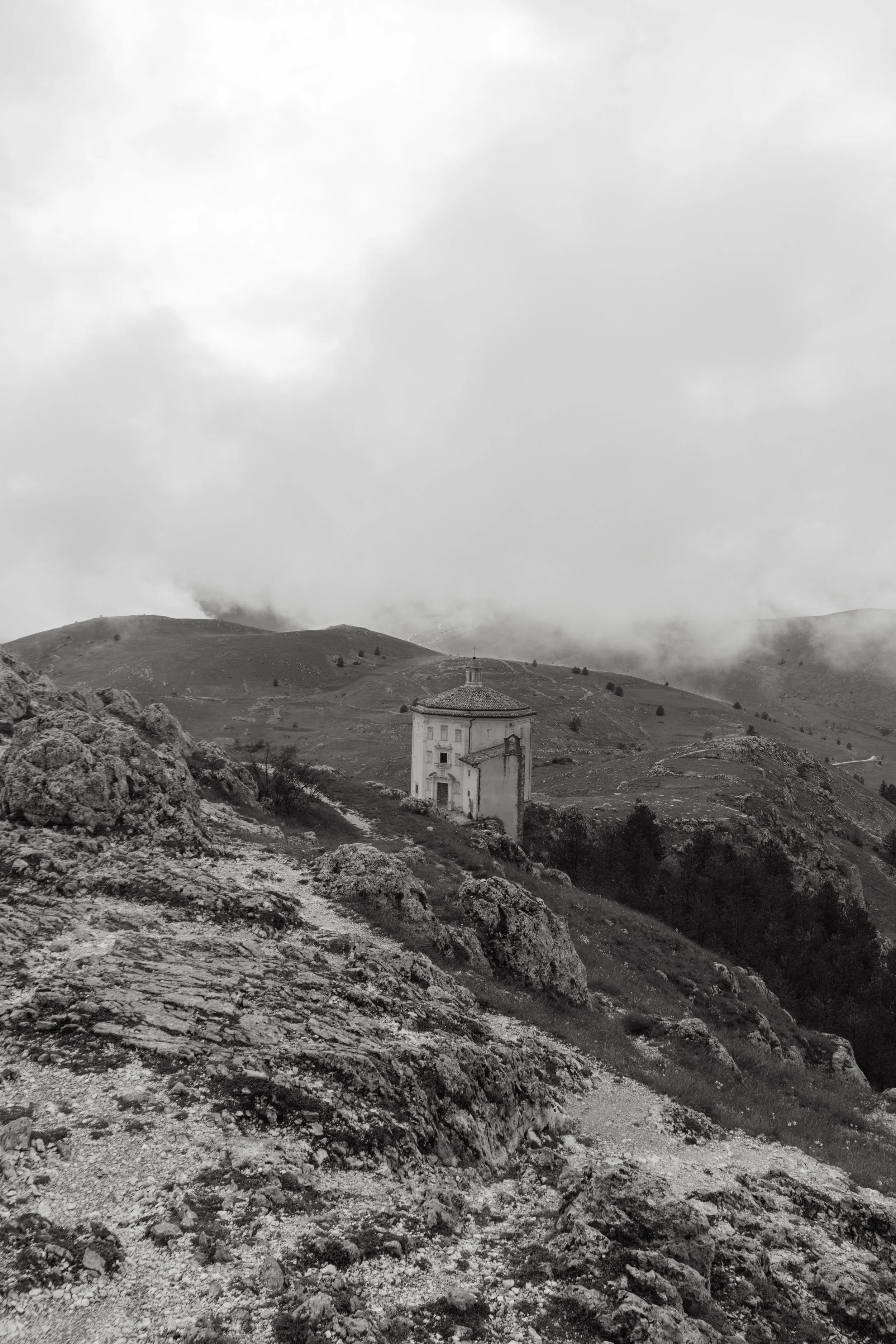 A black and white photo of a small building on a hillside, surrounded by rugged terrain and distant rolling hills under cloudy skies.