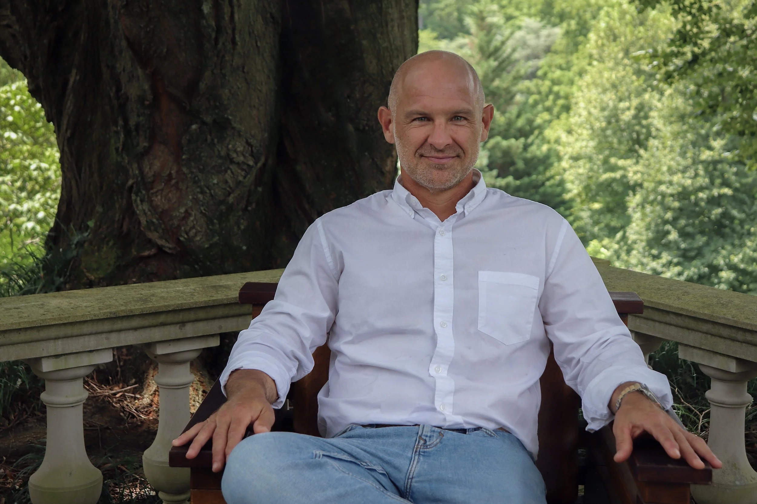 A handsome man sitting on a bench outdoors with greenery and a large tree in the background, wearing a white button-up shirt and jeans.