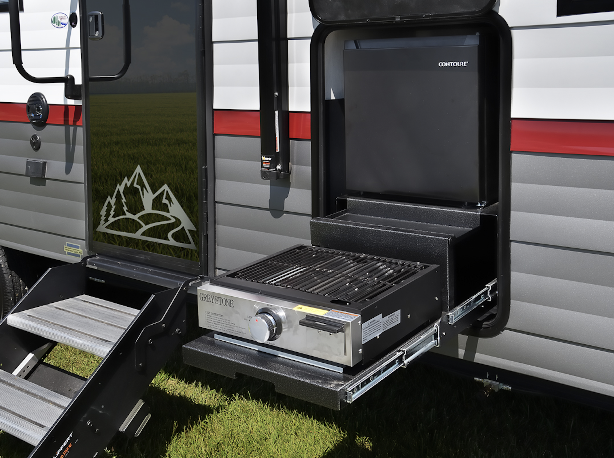 A portable outdoor camping kitchen setup with a small grill, a mini refrigerator, and stairs leading into a travel trailer. The trailer features a mountain graphic near the door and is situated on grass.