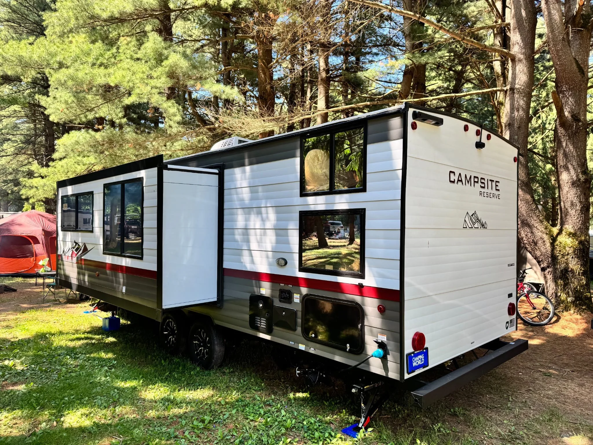 A white and gray camper trailer with black trim, parked on grassy ground under trees at a campground. It has multiple windows, and a slide-out section. There's a red tent and a bicycle in the background.