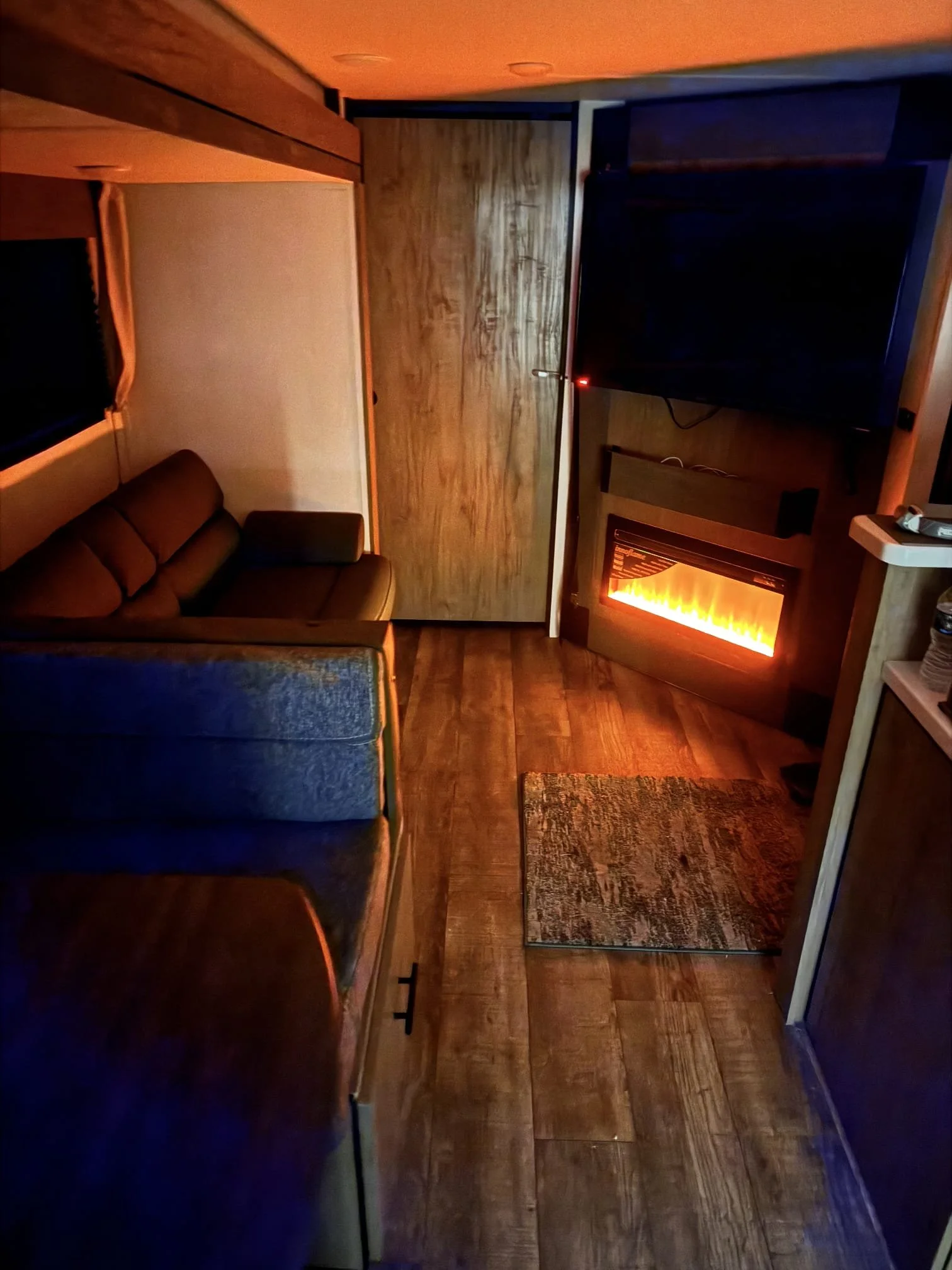 Cozy living room with a brown sofa, a large flat-screen TV, a small rug, a wall-mounted electric fireplace with orange glow, and wooden paneling on the walls.
