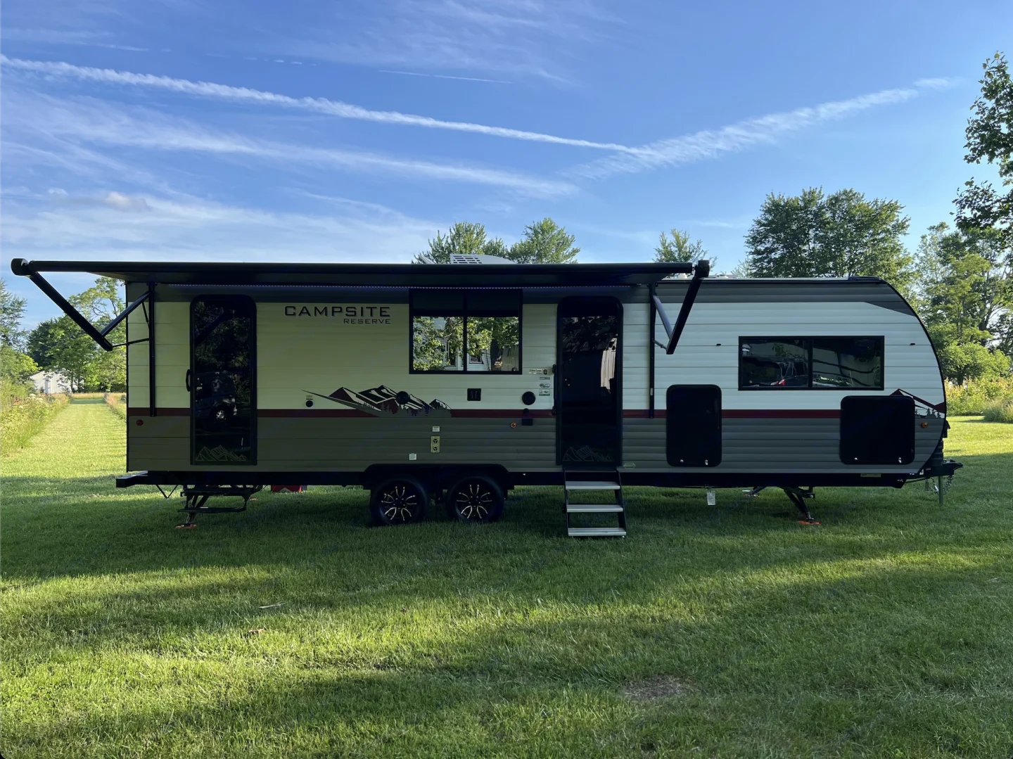 A white and gray travel trailer with a black logo that reads 'Campsite Reserve,' parked on a grassy area under a blue sky with wispy clouds and trees in the background.
