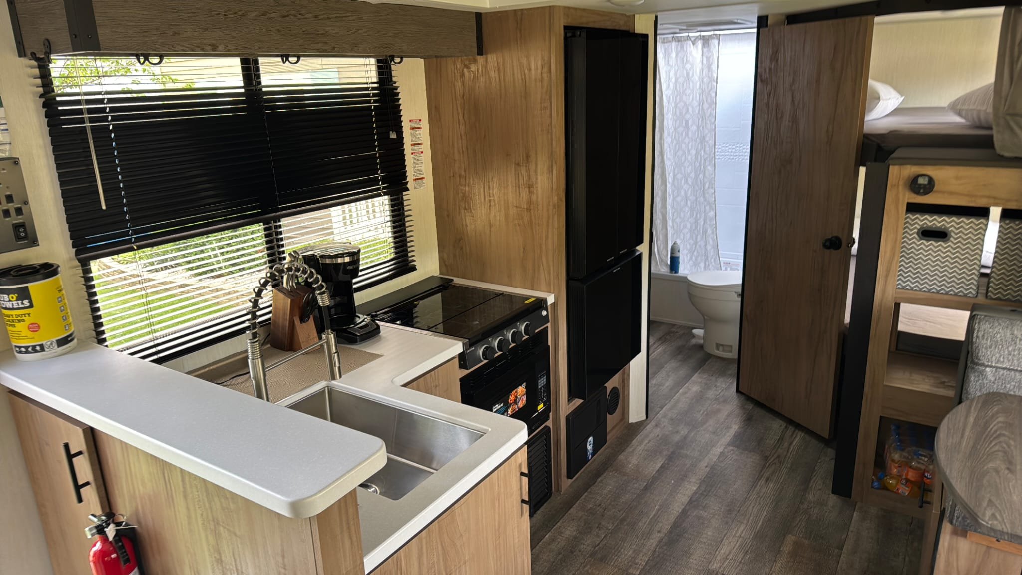 Interior of a compact kitchen and living space with a sink, coffee maker, stove, refrigerator, and a bathroom with a toilet visible through an open door.