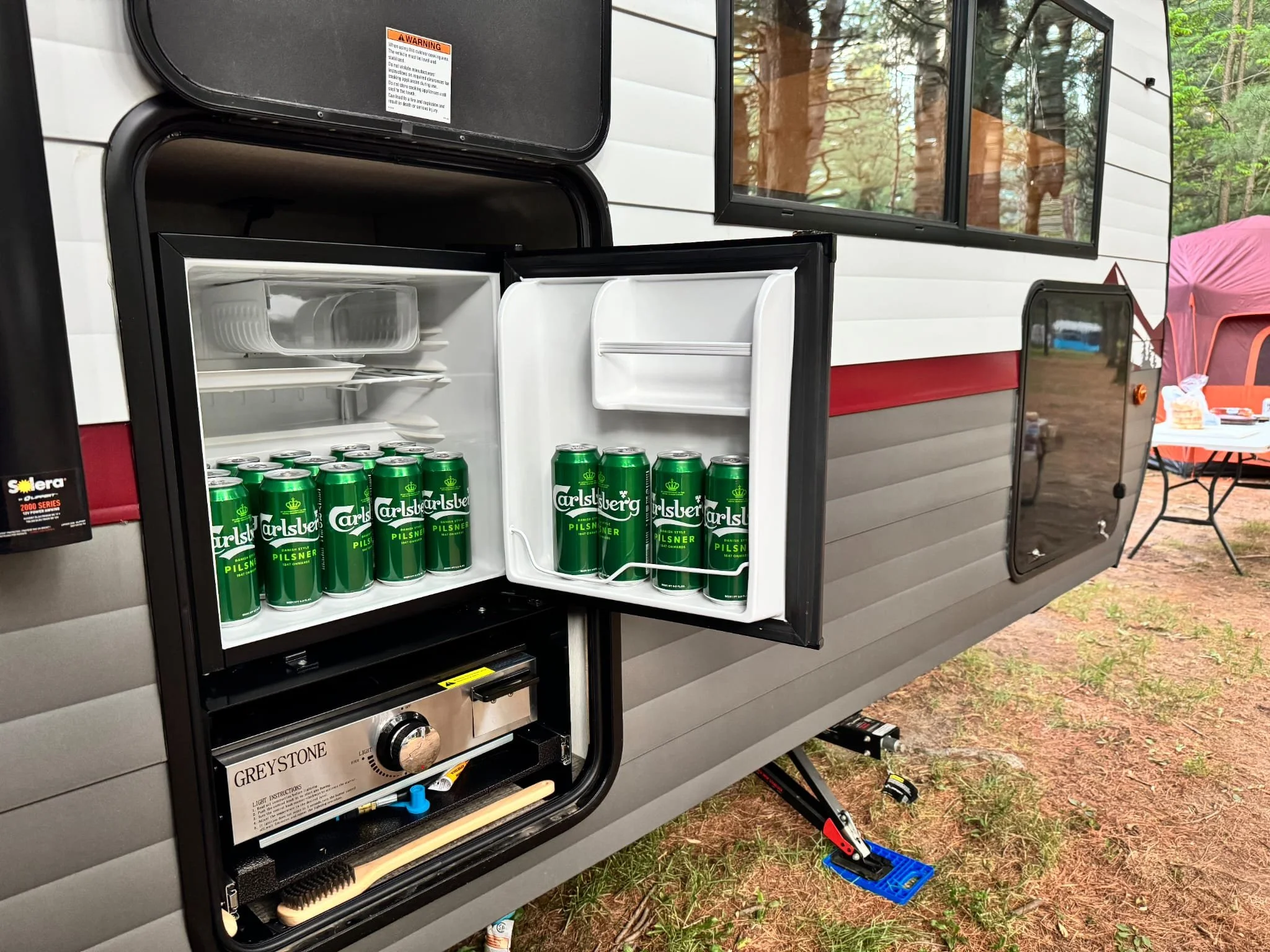 Small refrigerator inside a camper trailer, stocked with Carlsberg beer cans on the bottom shelf, with an outdoor camping setup visible in the background.