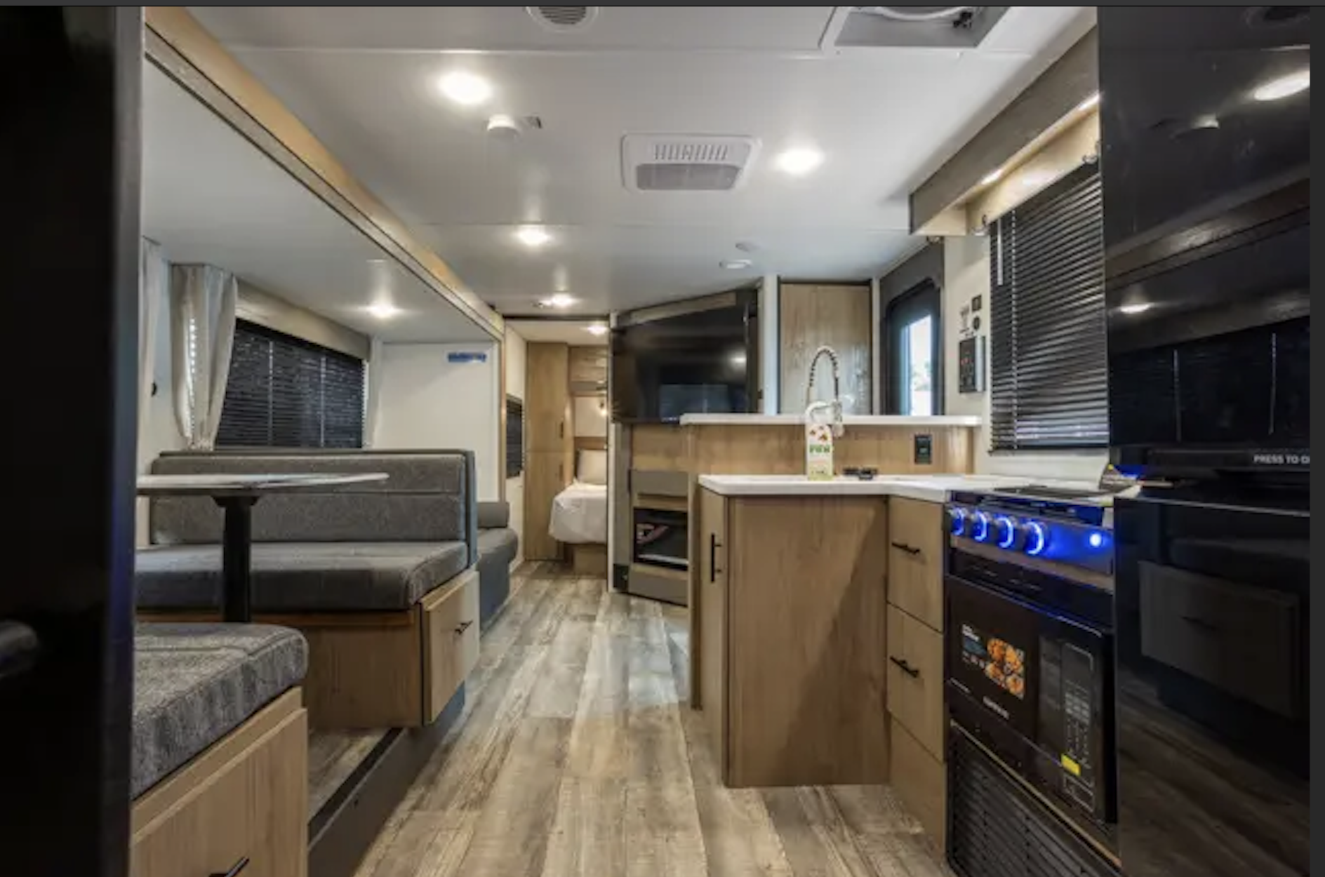 Interior of a modern RV with a small kitchen, a dining area with booth seating, a TV, and a bedroom in the background.