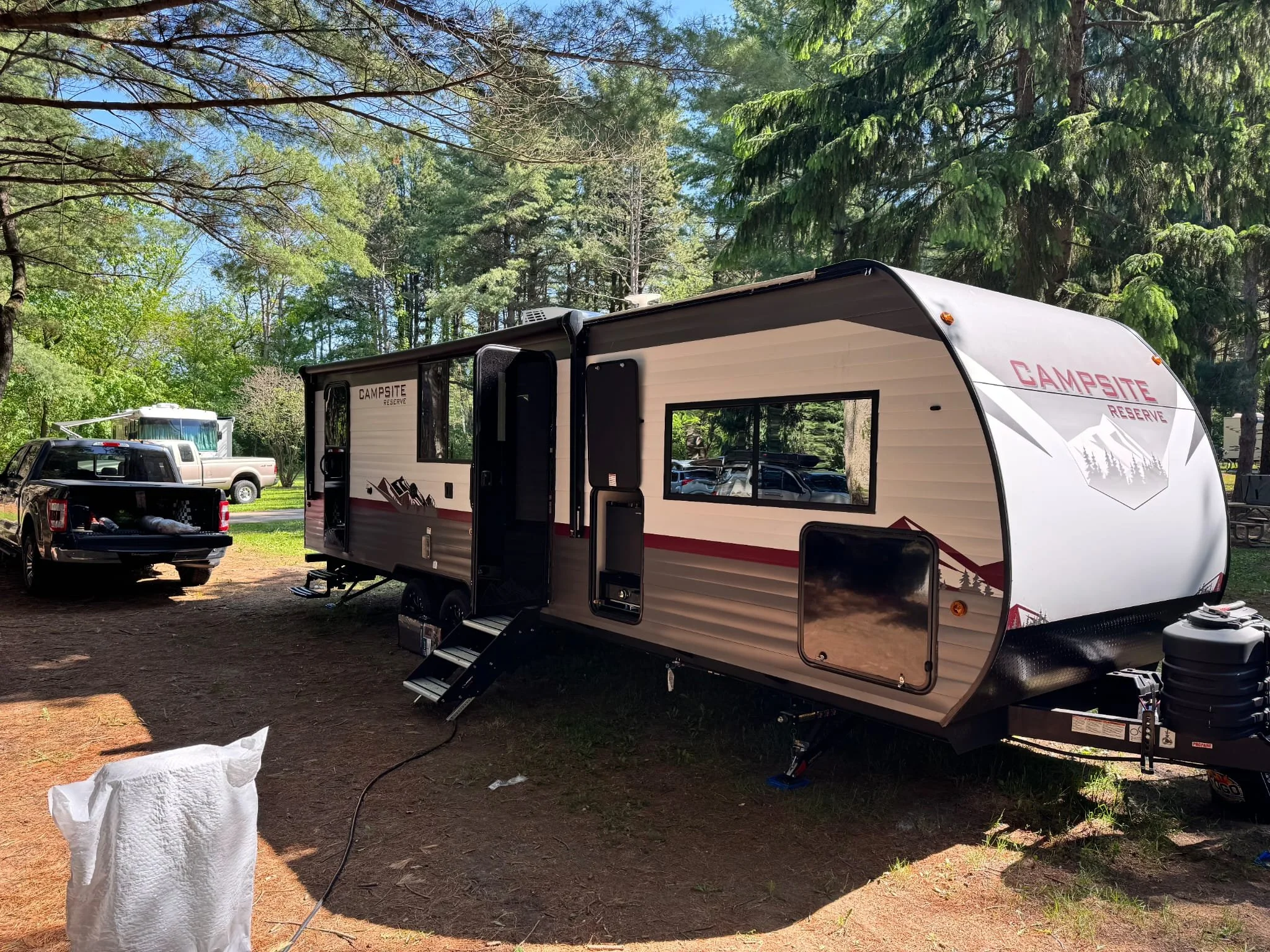 A large white and gray camping trailer parked in a wooded campsite with trees surrounding it, and a black pickup truck nearby.