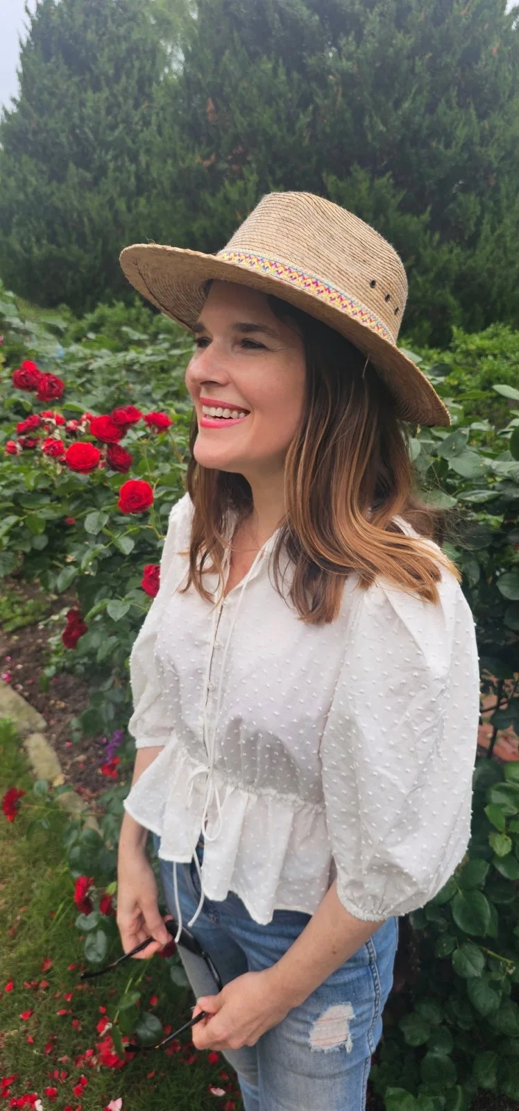 A woman smiling in a garden with green trees and blooming red roses, wearing a straw hat, white blouse, and ripped jeans.