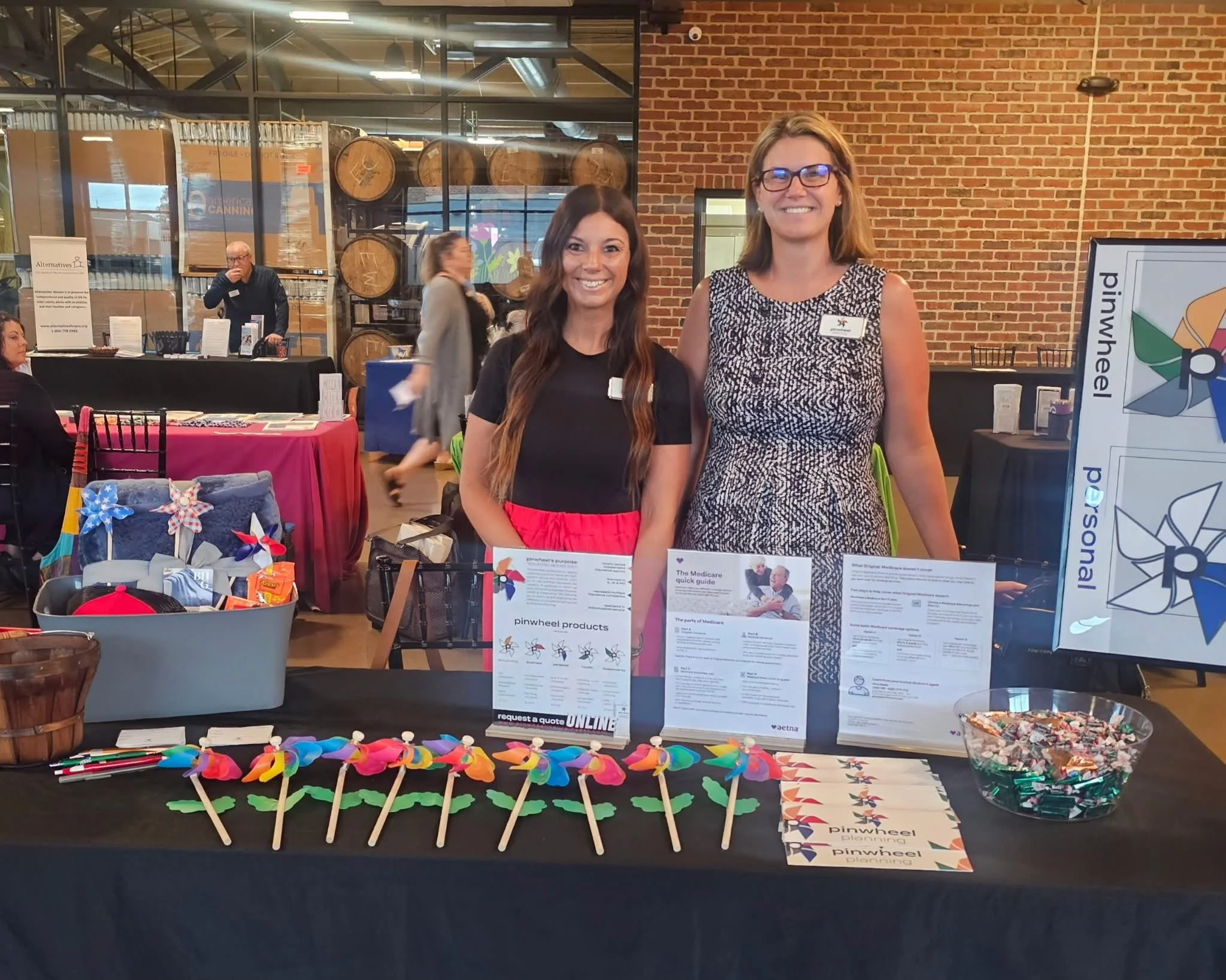 Two women standing behind a table at an indoor event, smiling for the camera, with pamphlets and colorful windmill decorations on the table, in front of a brick wall and wooden barrels.