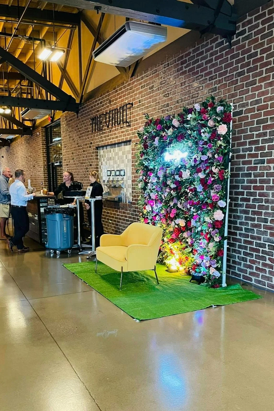 A yellow armchair on a green carpet in front of a floral backdrop with pink and purple flowers and green leaves, illuminated by colorful lights. The backdrop on a brick wall in the background, with a bar area and people standing nearby.