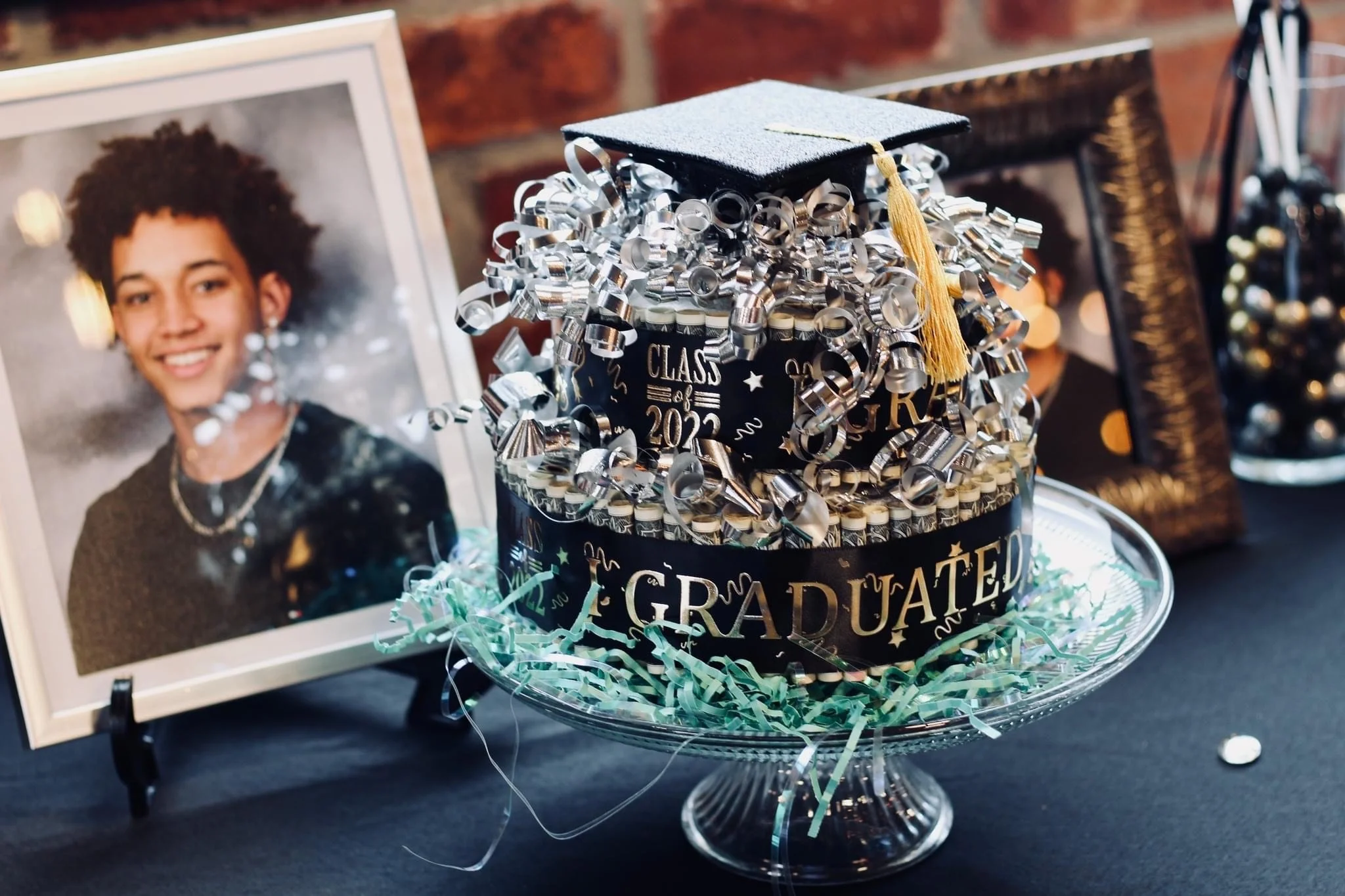 Graduation-themed graduation cap decorated with silver and black ribbons, placed on a glass stand. Nearby are framed photos of a smiling graduate with curly hair and a framed graduation picture. The display includes a black box with 'Class of 2022' written on it, surrounded by confetti and shredded paper for decoration.