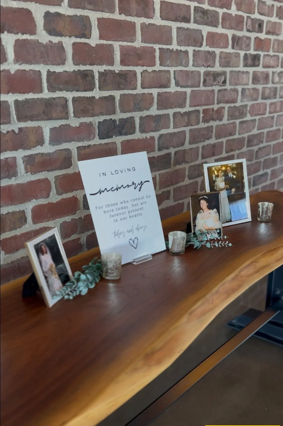 A memorial display on a wooden table against a brick wall, featuring framed photographs of a couple, a framed photo of a woman in a white dress, a sign with a condolence message, and small candles with decorative greenery.