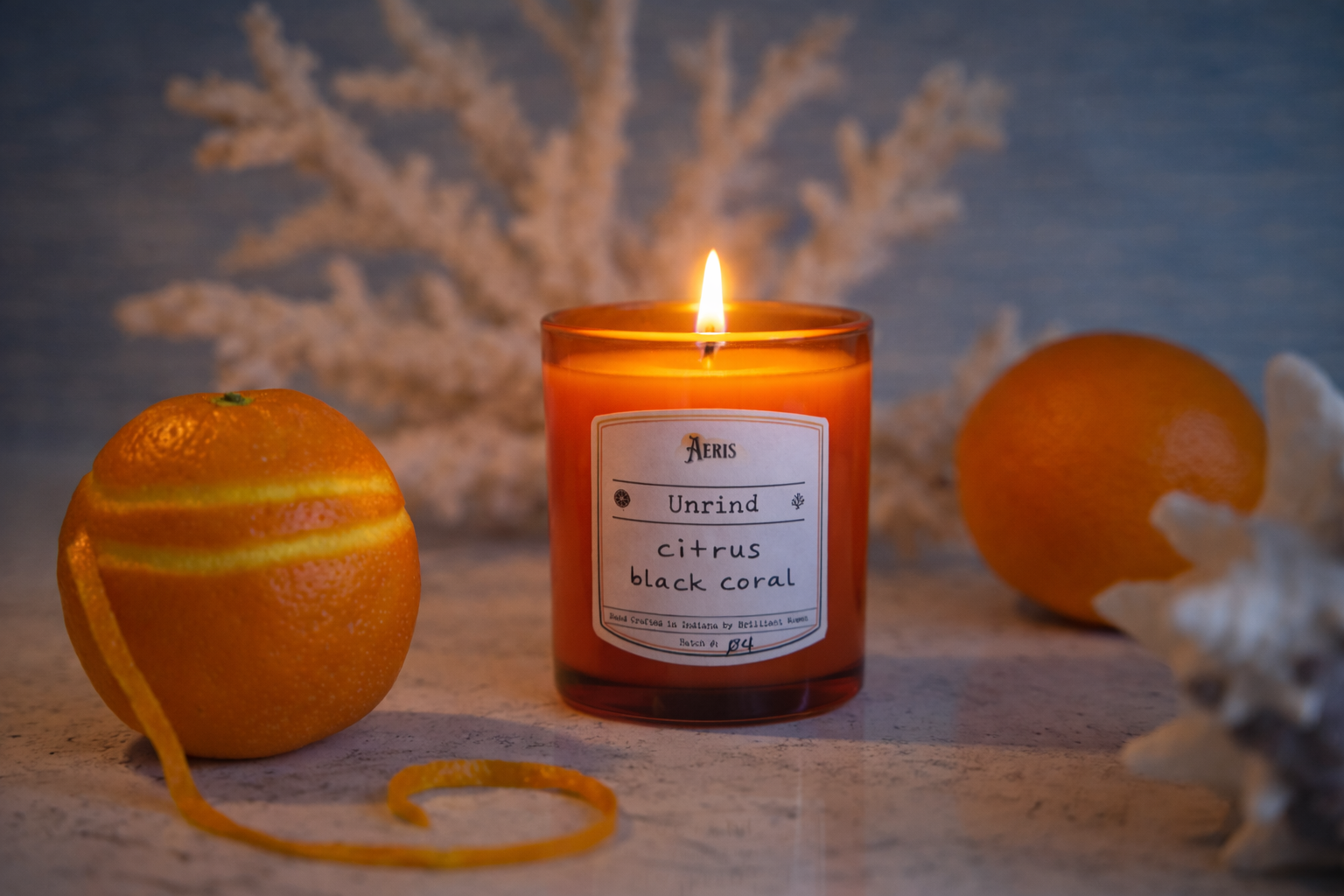 A lit amber glass candle labeled “Unrind” sits on a quartz countertop, flanked by fresh oranges and curling citrus peel, with soft coastal textures blurred in the background.
