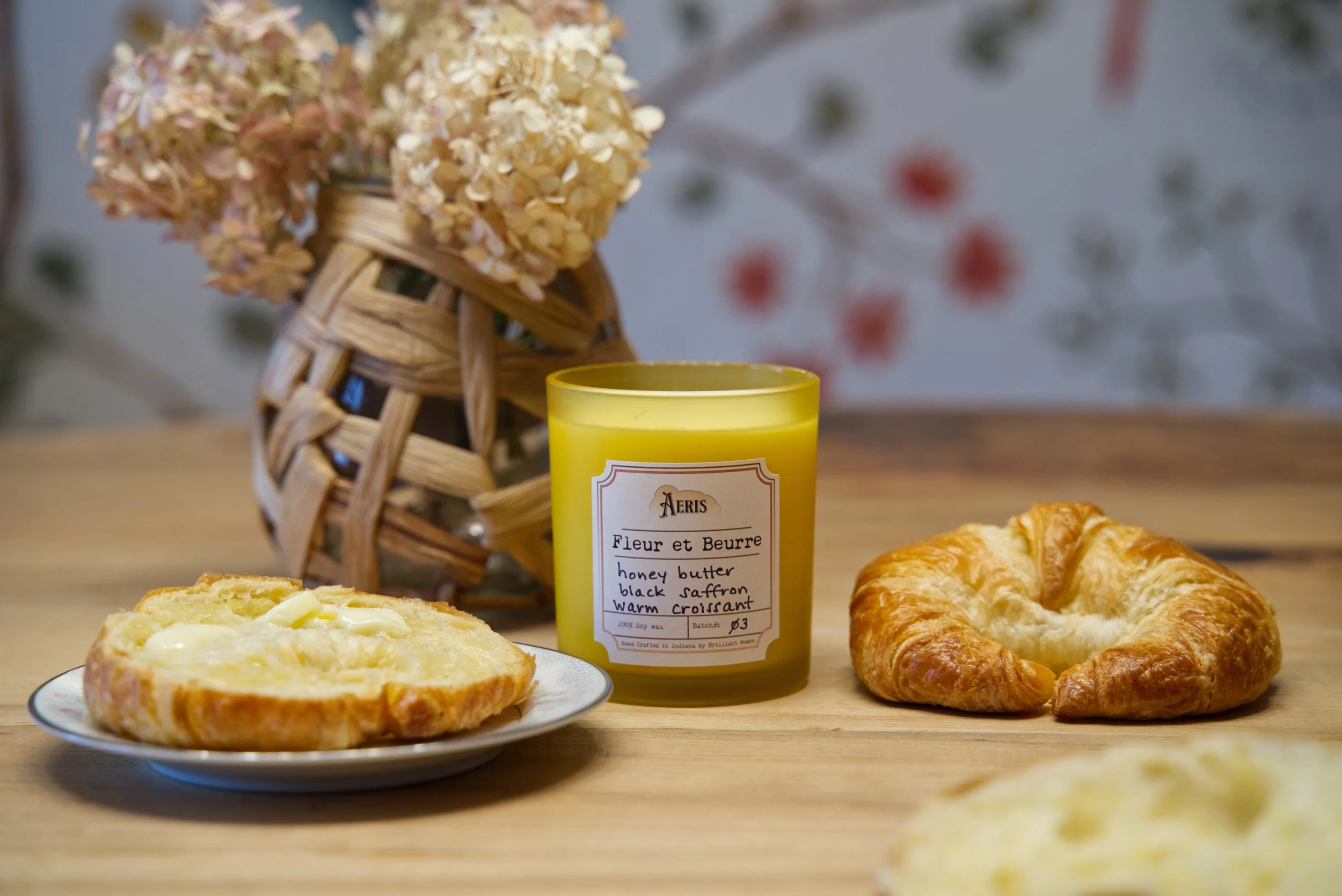 Aeris Fleur et Beurre candle in yellow glass beside croissants and buttered toast on wooden table with floral background