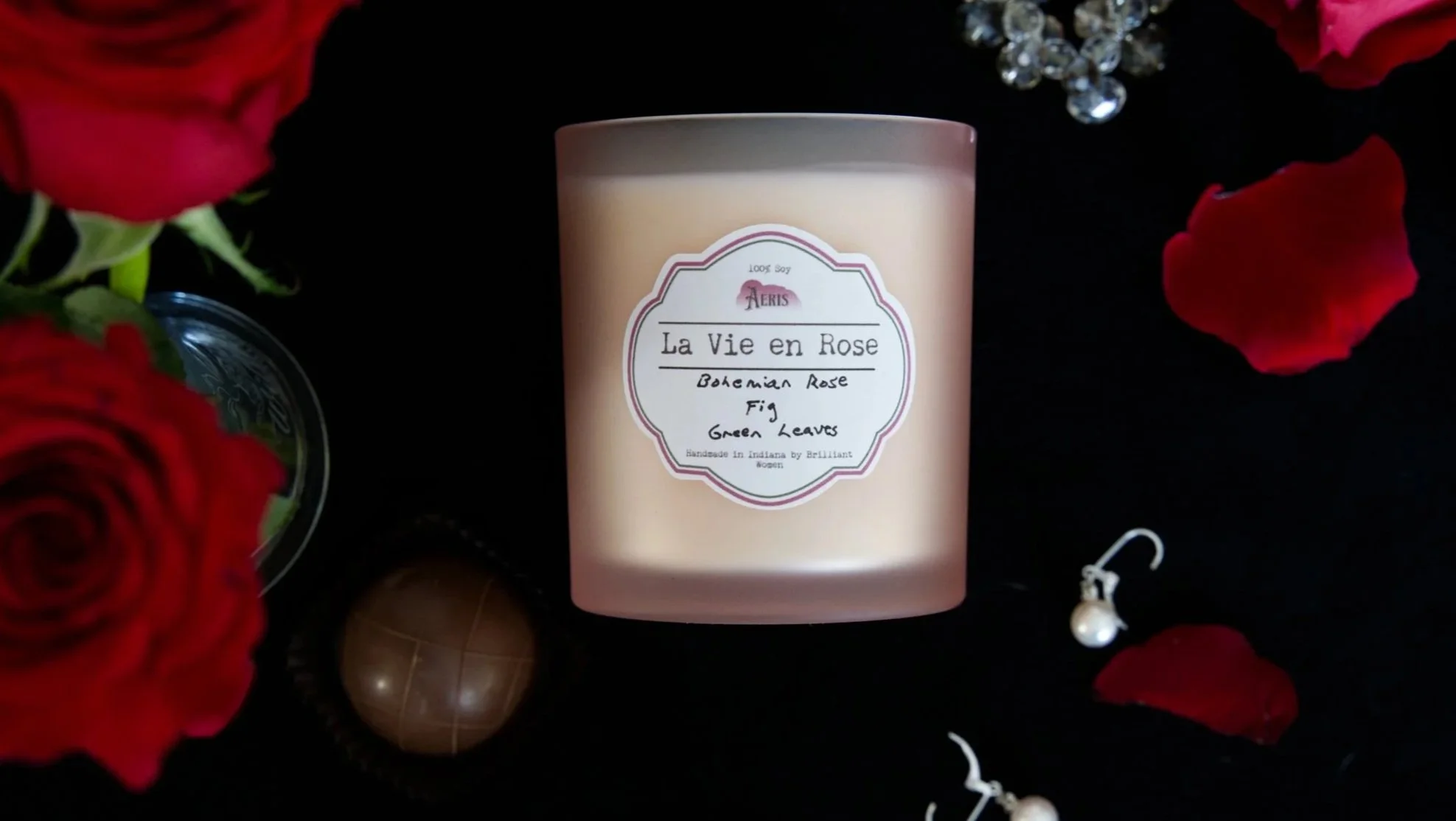 Aeris La Vie en Rose candle in a blush glass vessel, styled with red roses, scattered petals, pearls, and jewelry on a black background.