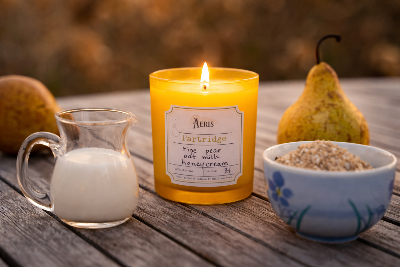 A lit amber glass candle labeled “Partridge” sits on a weathered wooden table, surrounded by ripe pears, a small pitcher of milk, and a bowl of oats, evoking a warm, rustic autumn setting with soft golden light.