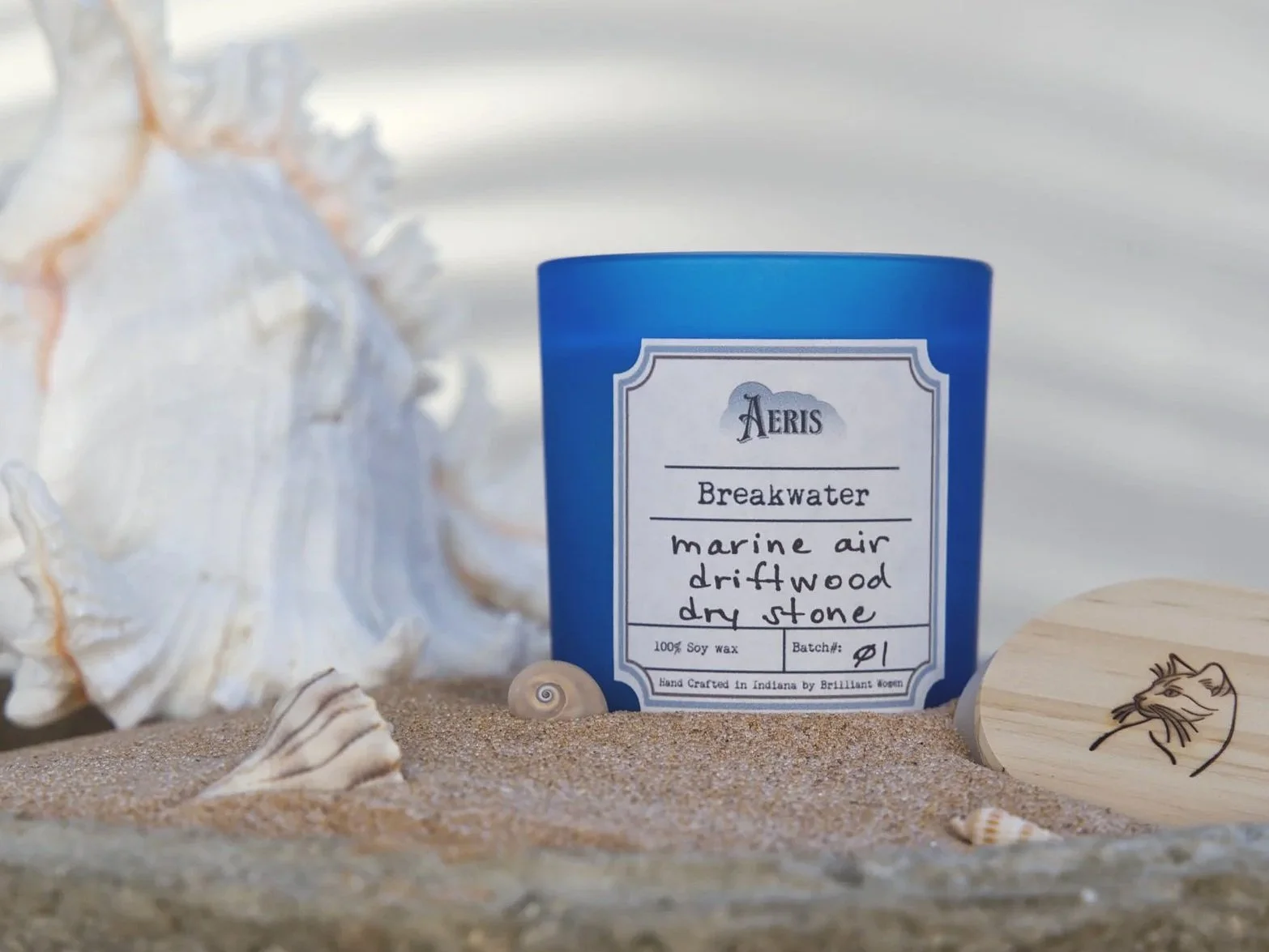 Aeris Breakwater candle in a blue glass vessel styled in sand with seashells and a large conch shell, evoking a coastal marine atmosphere.
