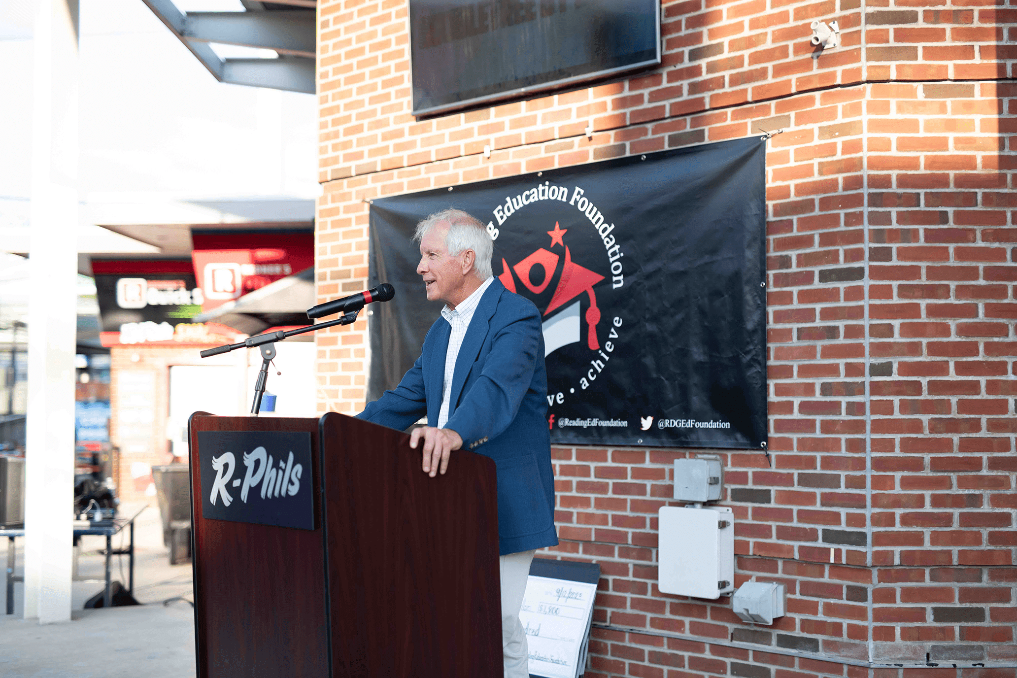 An elderly man in a blue suit and white shirt speaking at a podium outdoors, with a black banner behind him that reads 'Racing Ed Foundation,' attached to a brick wall.