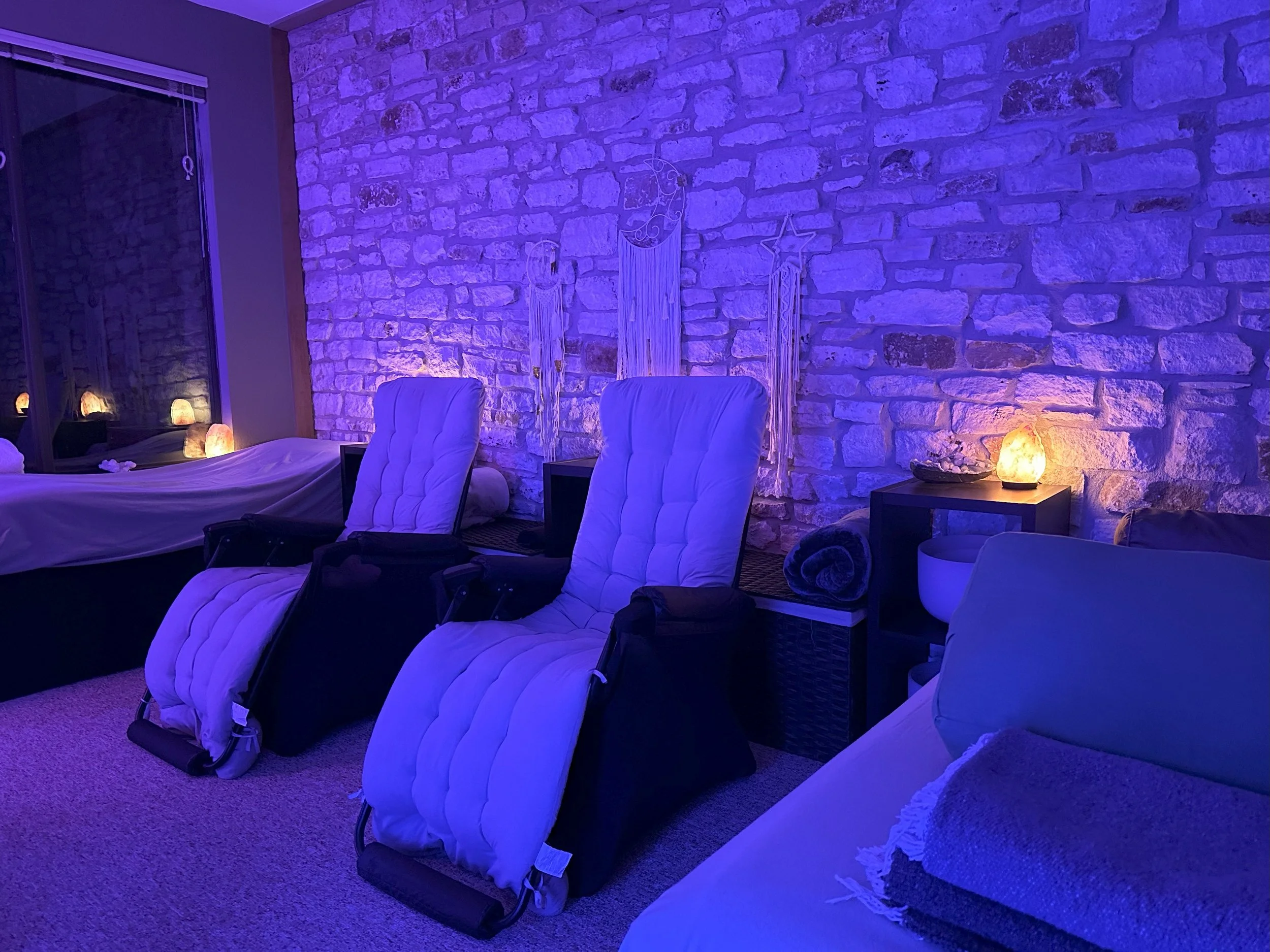 A cozy room with two massage chairs illuminated by soft purple and warm lighting. The room has a stone accent wall decorated with hanging dreamcatchers. To the left, there's a bed with salt lamps on a side table, and at the right, a lounge chair with