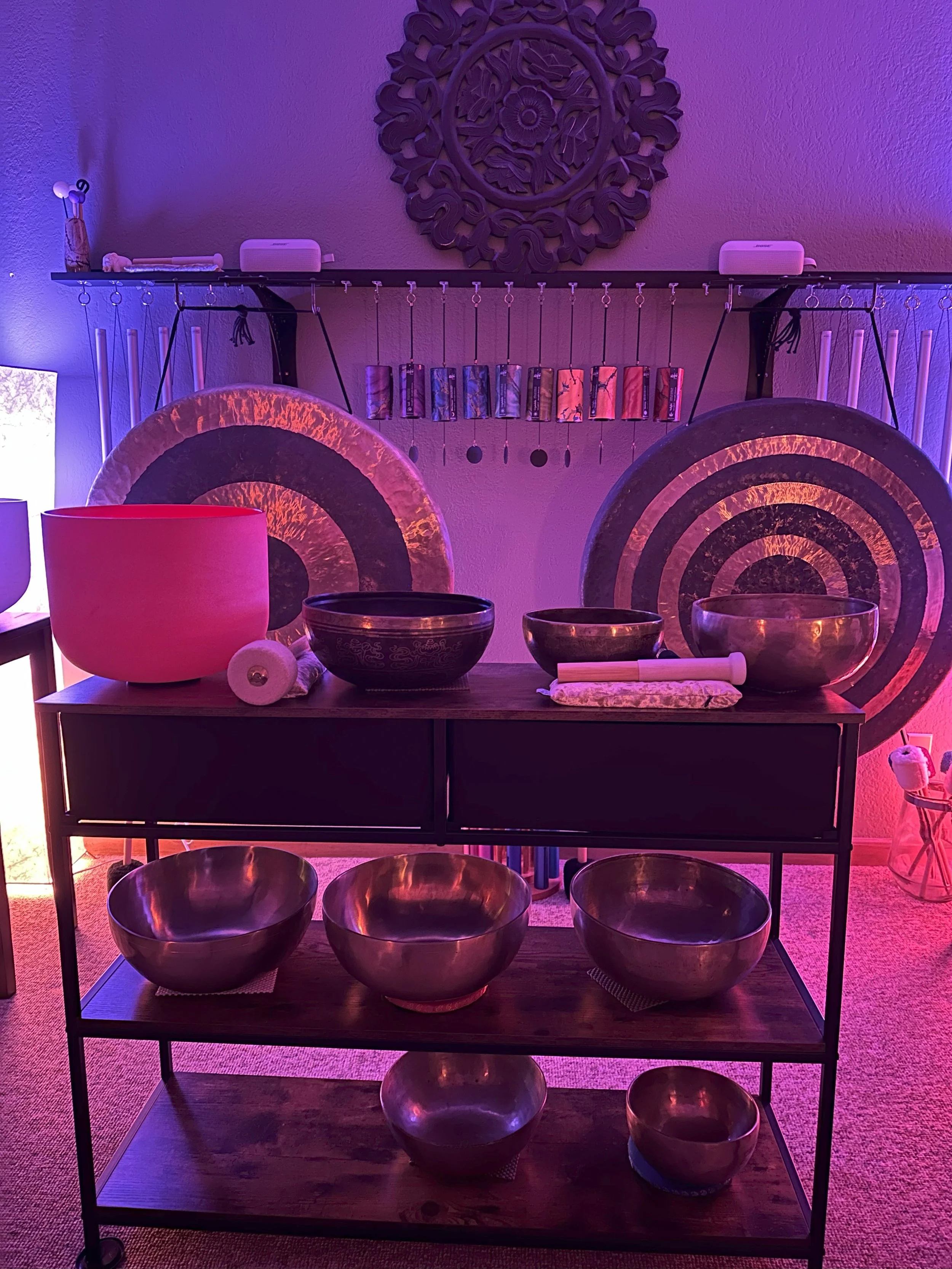 A collection of singing bowls and large gongs and wood shelving unit under purple lighting, with a wall hanging and additional items in the background.