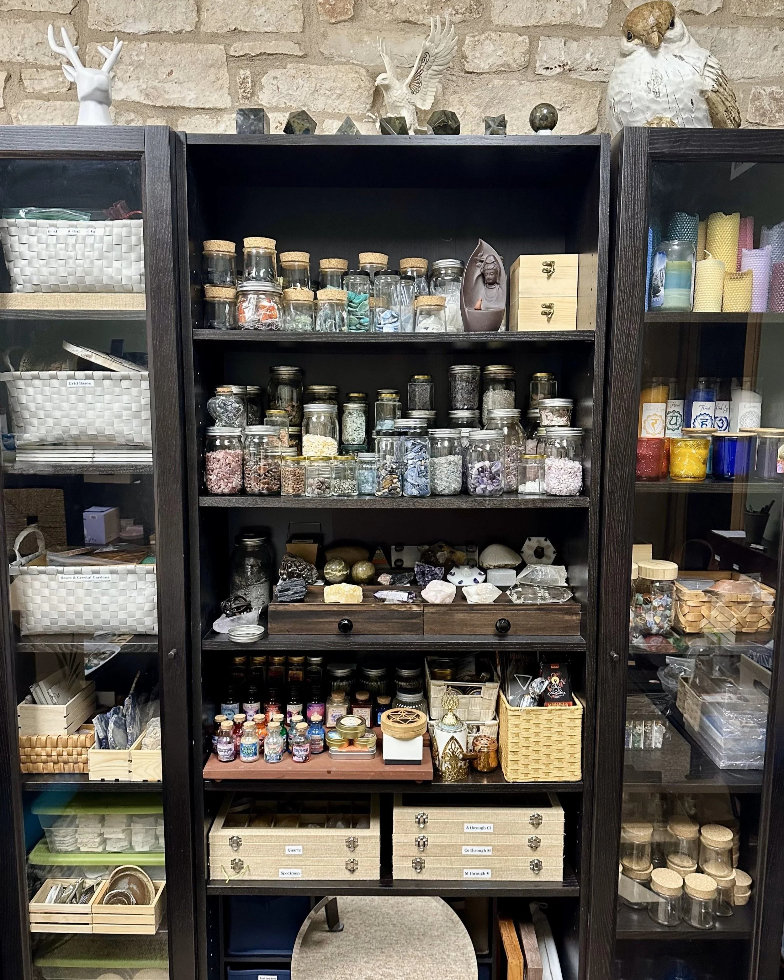 Shelf filled with glass jars containing crystals, herbs, and powders; decorative figurines on top, including an owl and a deer head