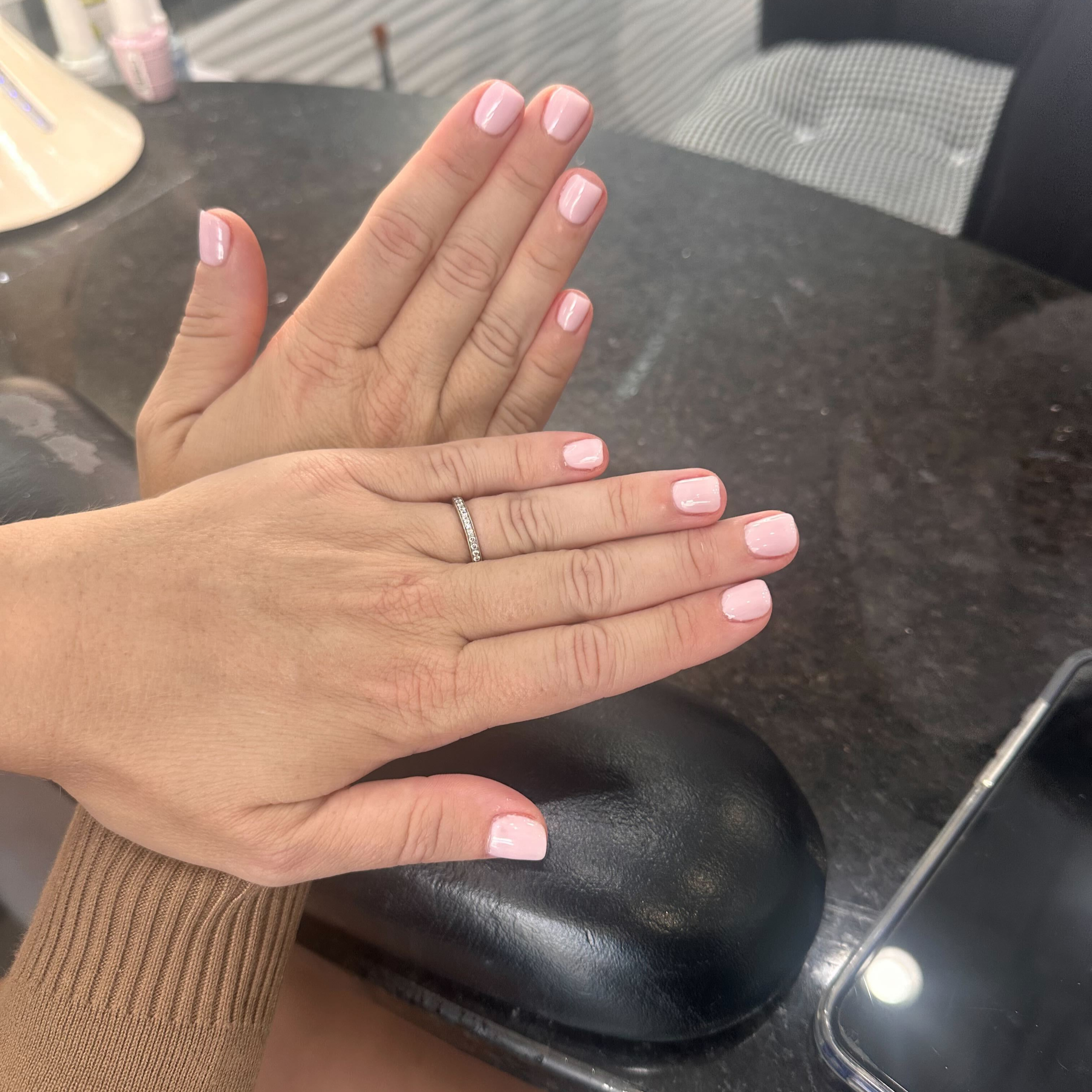 Close-up of a person showing their hand with freshly painted light pink nails, with a wedding ring on the ring finger, resting on an object, possibly a desk or counter, with a black surface and a metallic edge.