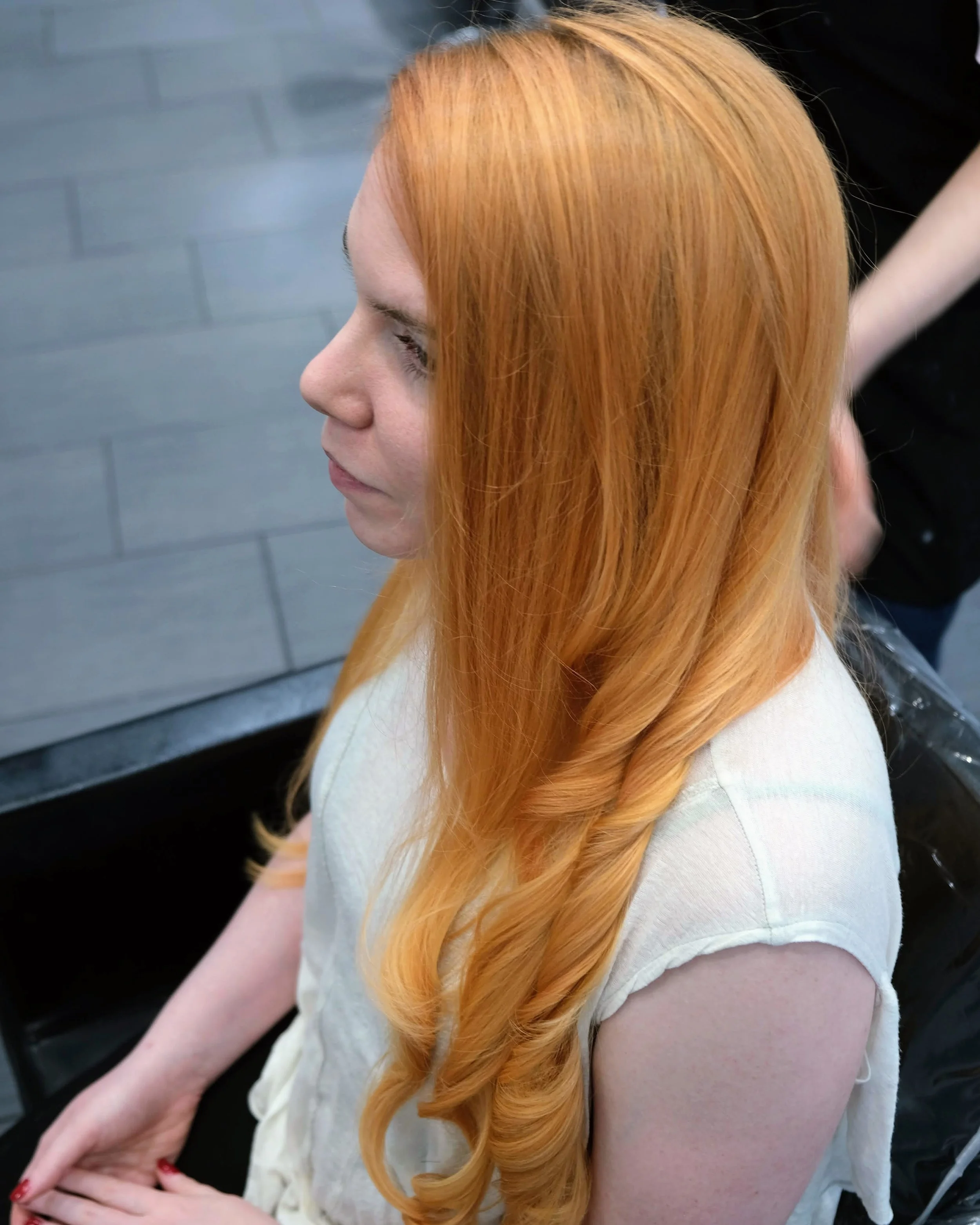 A woman with long, wavy, red hair sitting in a salon chair, wearing a white sleeveless top.